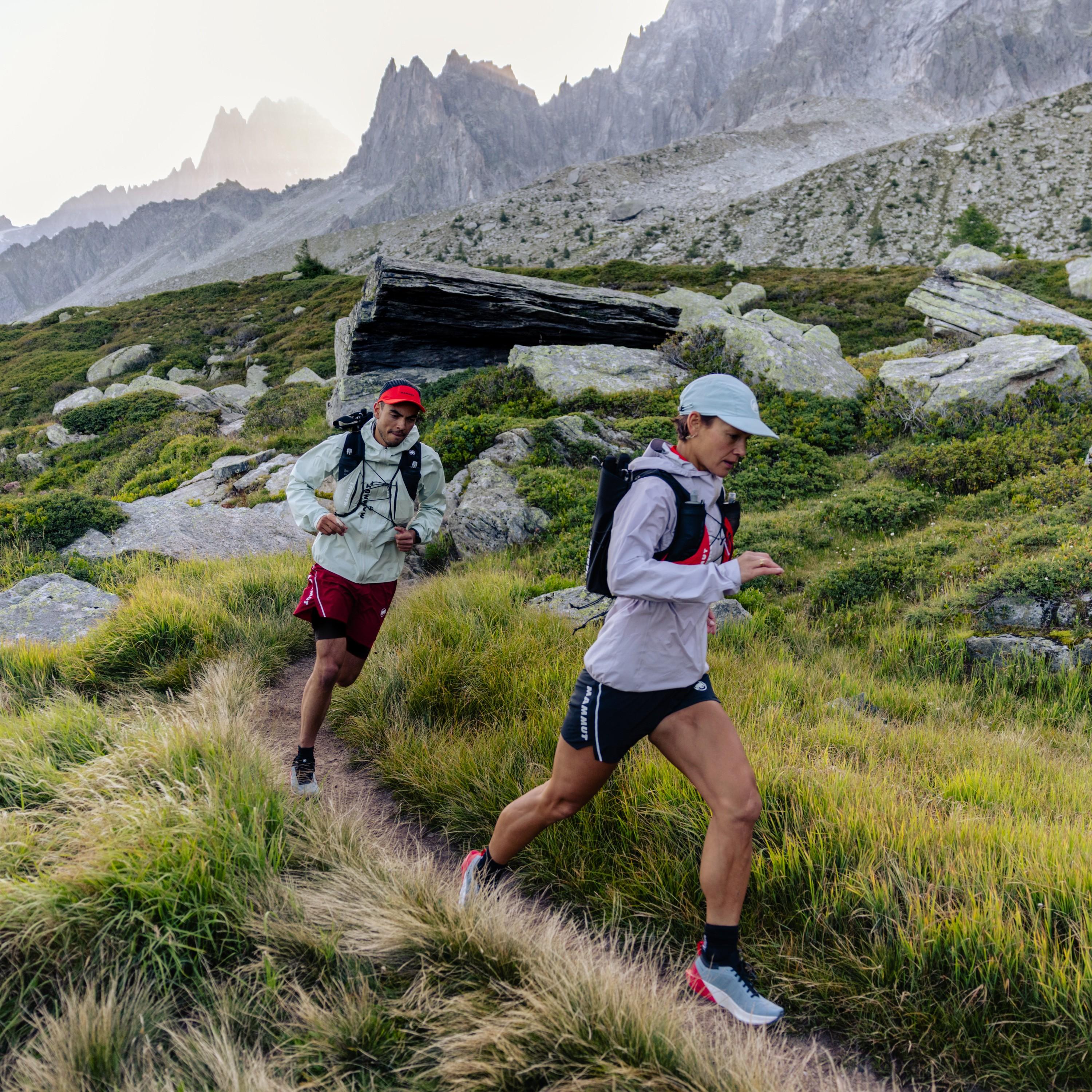 Two athletes in Mammut gear trail running on a grassy mountain path, rocky cliffs and alpine peaks rising in the background.