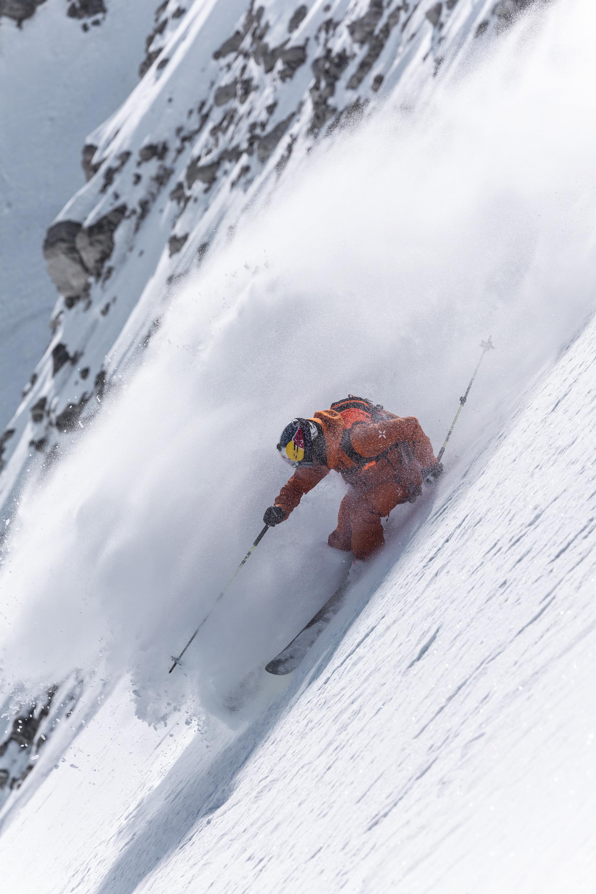 Skier in orange Mammut gear expertly maneuvers through deep snow on a steep mountain slope, creating a spray of powder.
