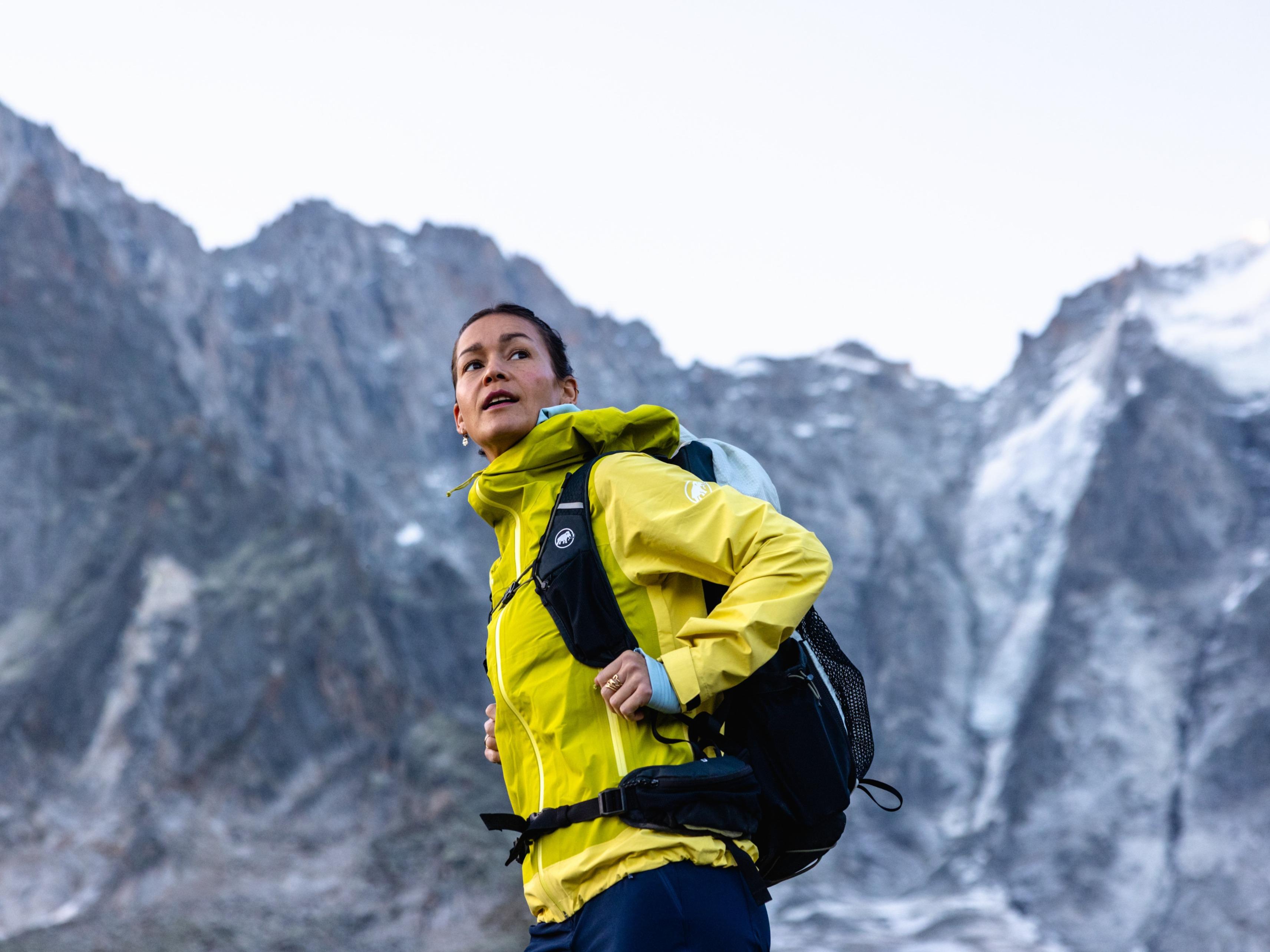 A woman in a white Mammut jacket hikes on a rocky mountain trail, with alpine lakes and rugged peaks visible in the scenic background.