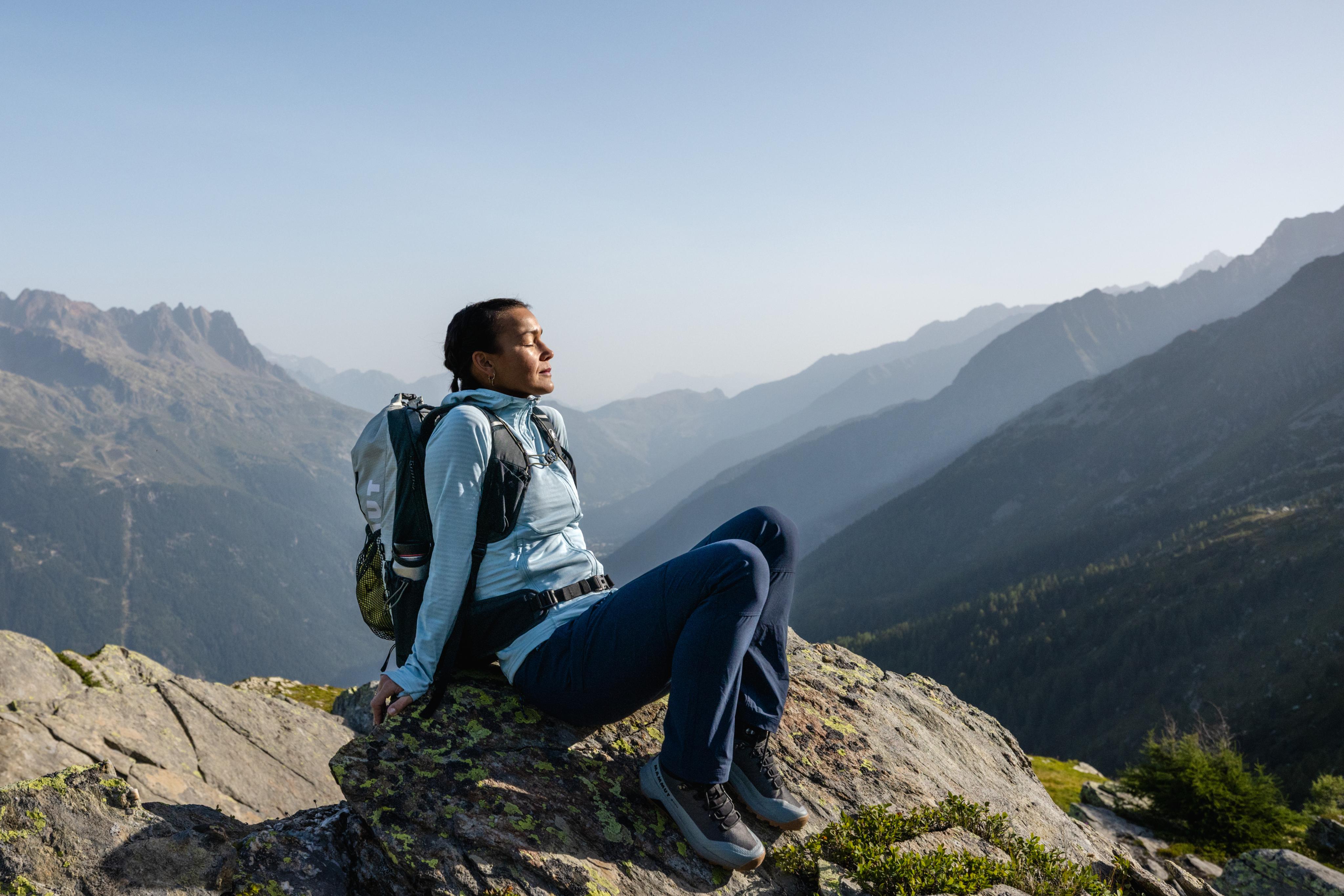 Woman in a yellow Mammut jacket hiking with a technical backpack before towering snow-covered mountain peaks.
