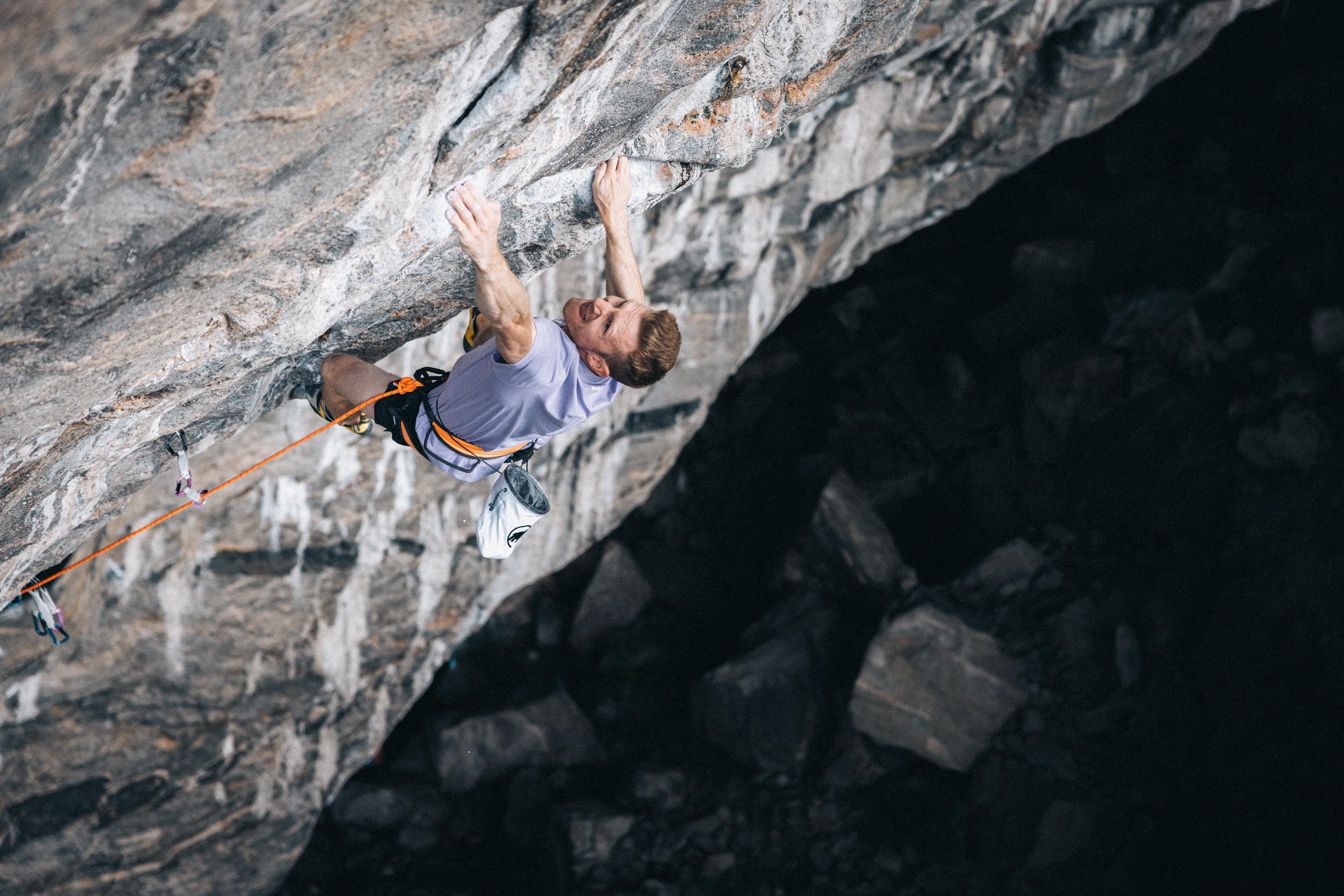 A climber wearing Mammut gear scales a steep rock face, gripping an overhang with rugged rocky terrain visible far below.