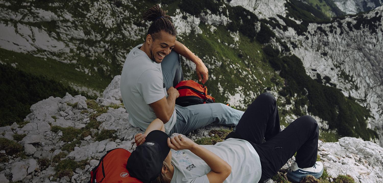 Two hikers wearing Mammut gear sit and laugh together on a rocky mountain ledge with Mammut backpacks, enjoying the scenic alpine view.