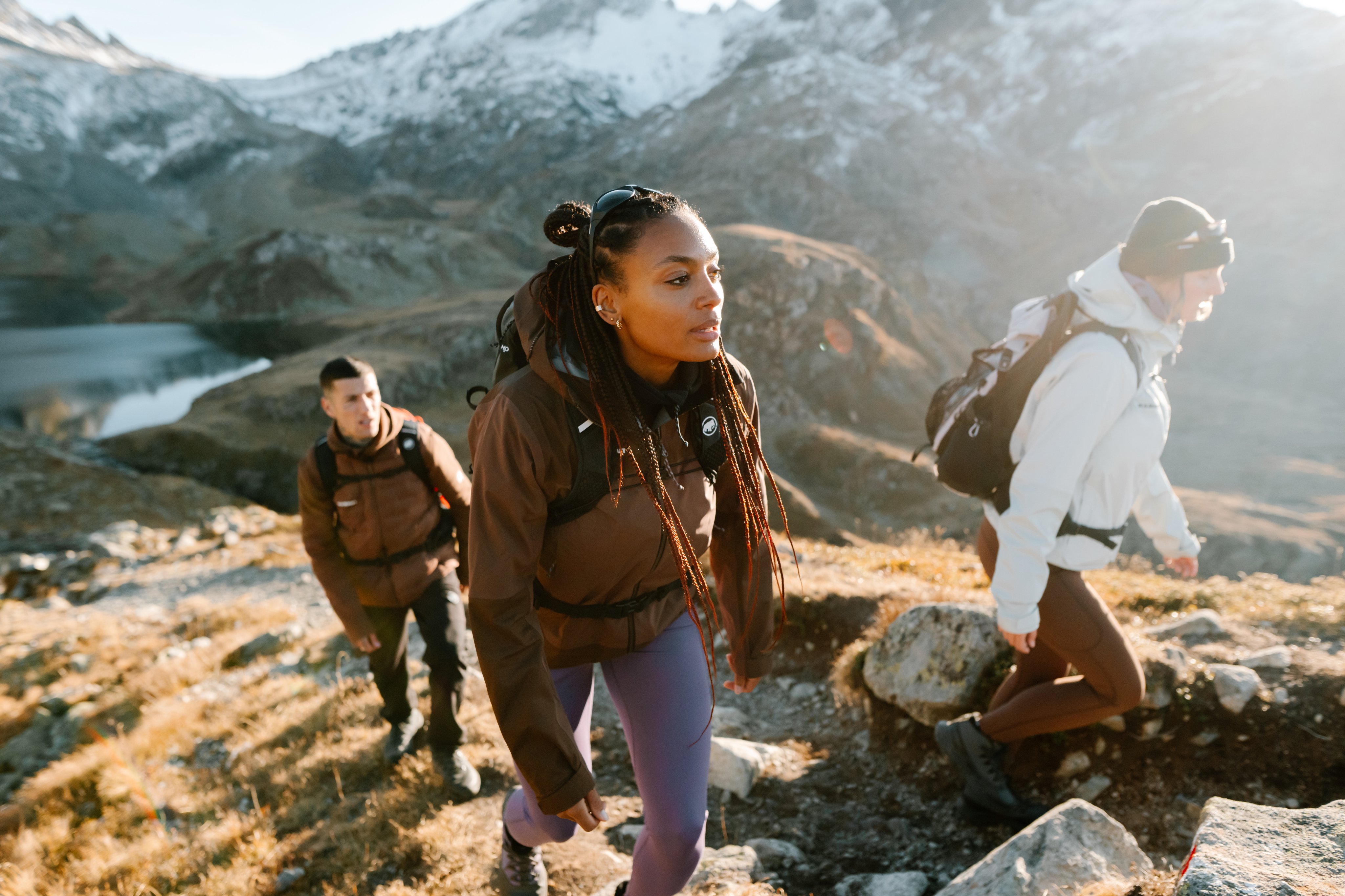 Three hikers in Mammut gear trek a rocky mountain trail, snowy peaks and a clear alpine lake visible in the background.