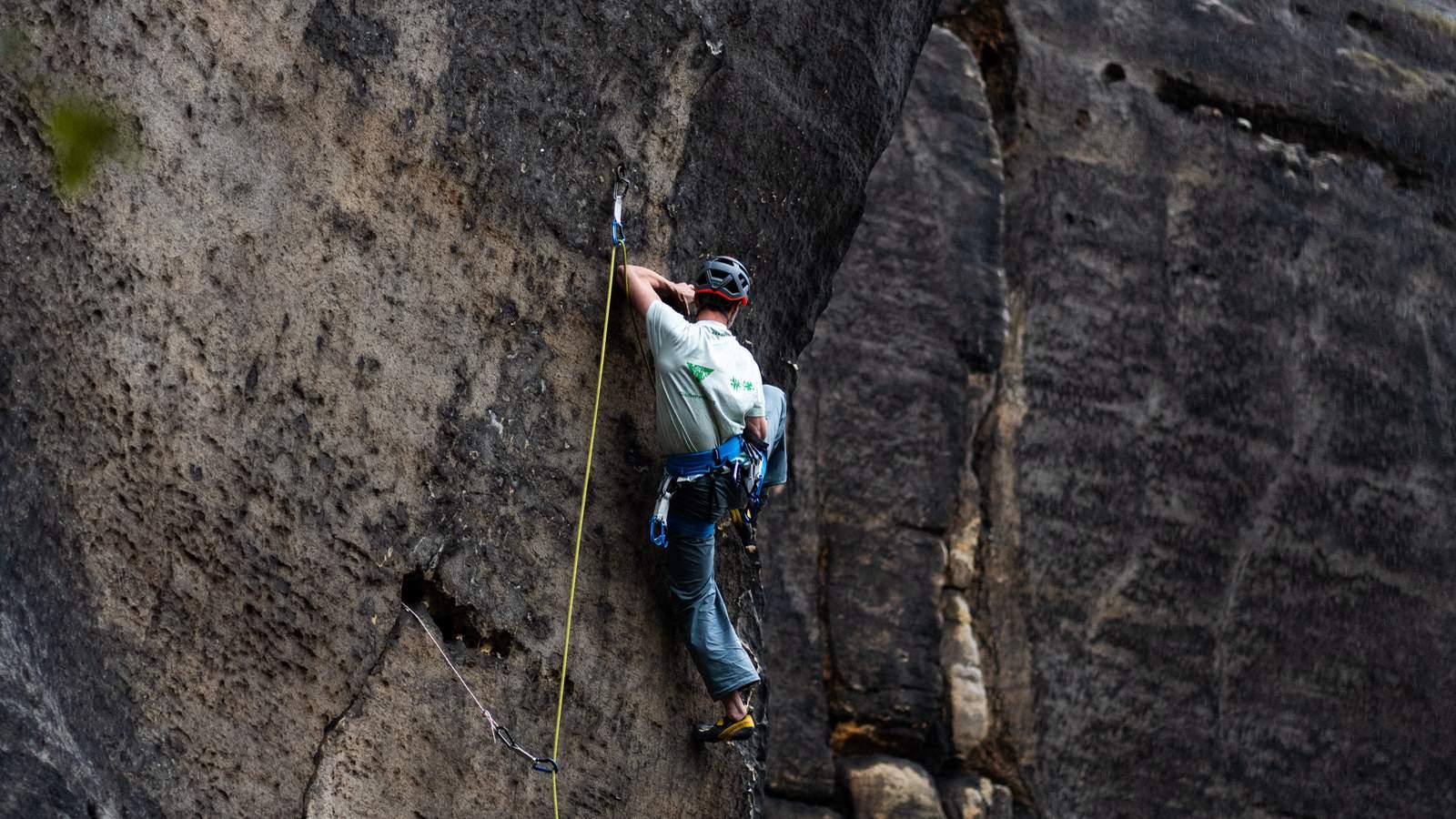 A climber in a Mammut helmet grips a rugged rock face one-handed, high above a dense forest landscape.