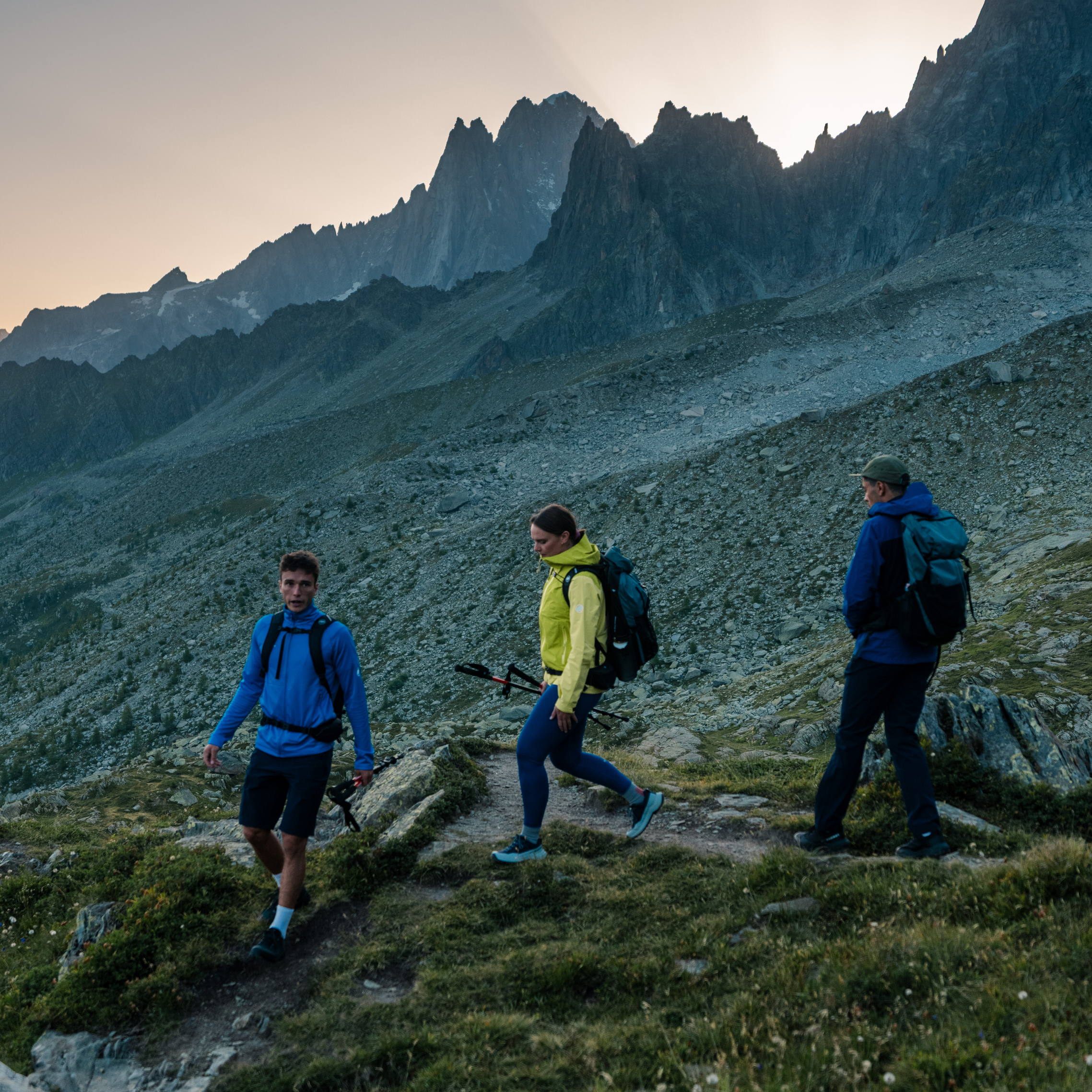 A hiker in a Mammut light jacket and distinctive purple pants ascends rocky terrain, with majestic mountains in the background under a clear sky.