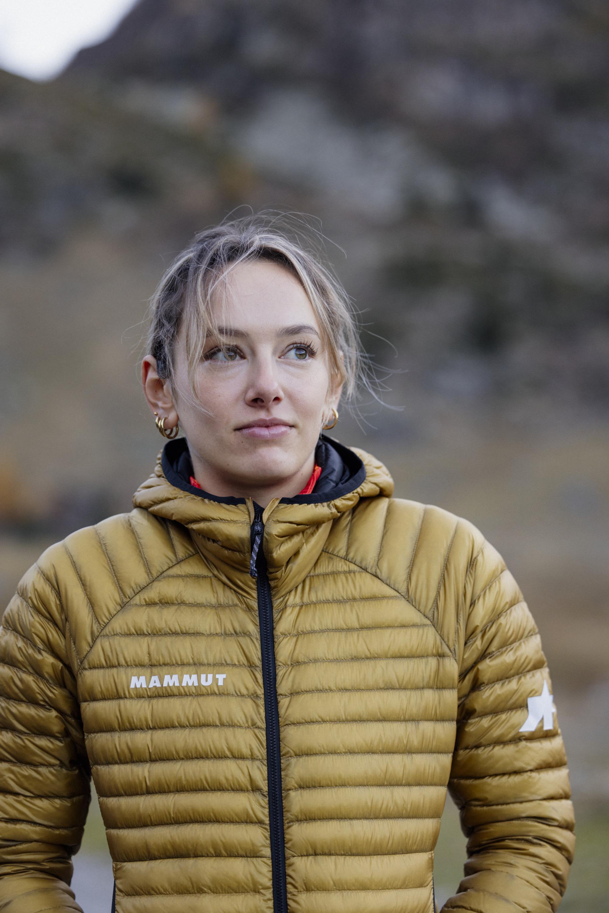 Person wearing a brown Mammut jacket, standing outdoors with a blurred natural mountain landscape in the background.