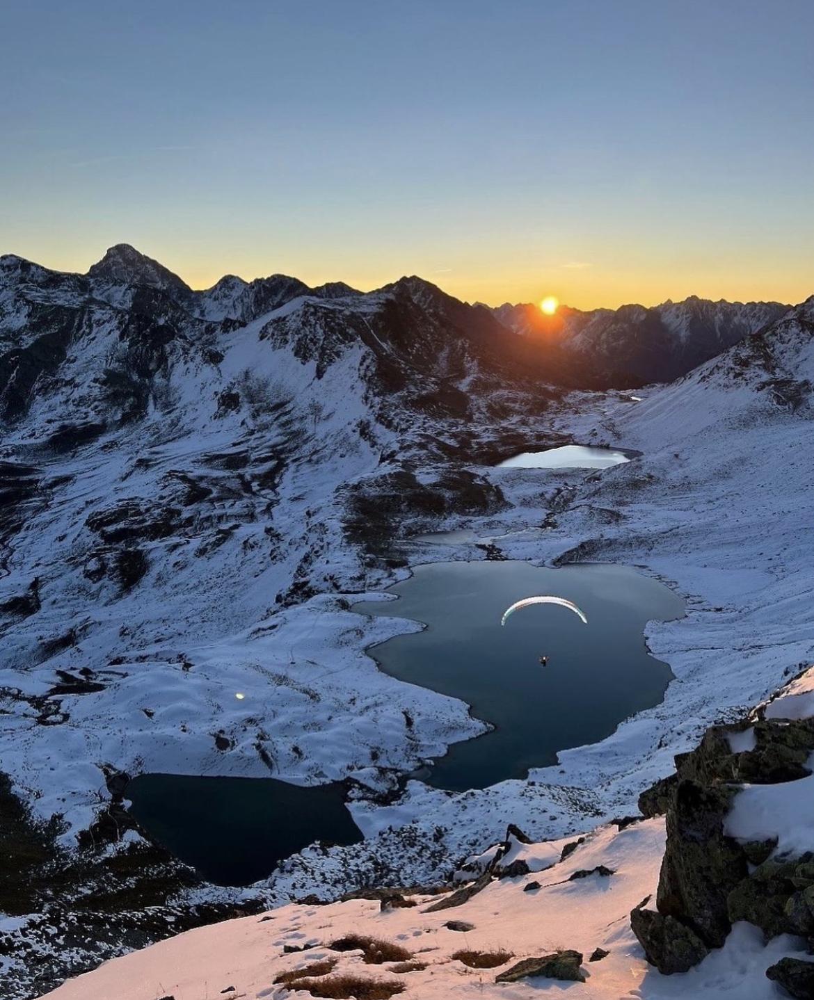 Mammut paraglider soaring over a snowy alpine mountain valley at sunset, with glacial lakes and rugged peaks in the background.