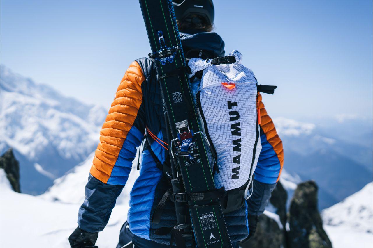 A skier in Mammut winter gear climbs a snowy mountain, carrying skis on their back and using an ice axe for mountaineering.
