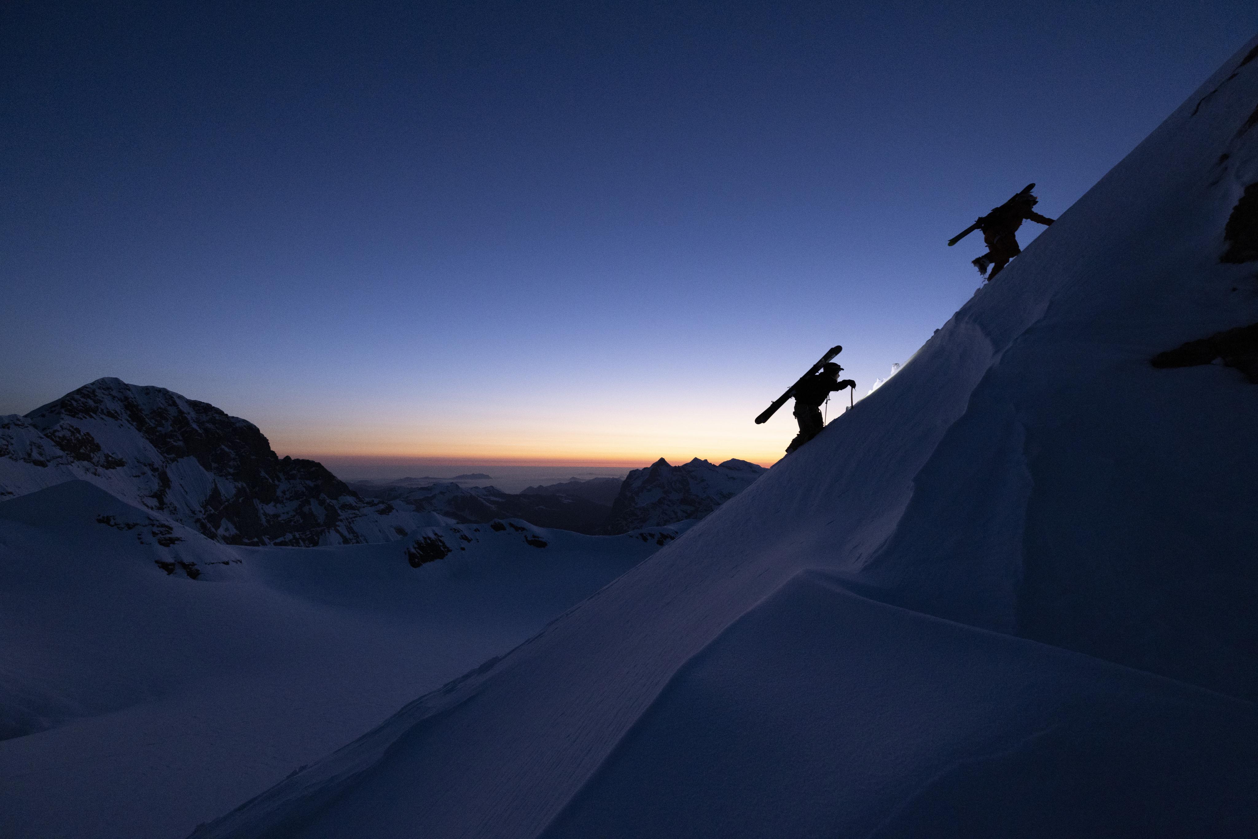 Two skiers, equipped with Mammut gear, hike up a snowy mountain at dawn, carrying their skis, with a silhouetted mountain range in the background.