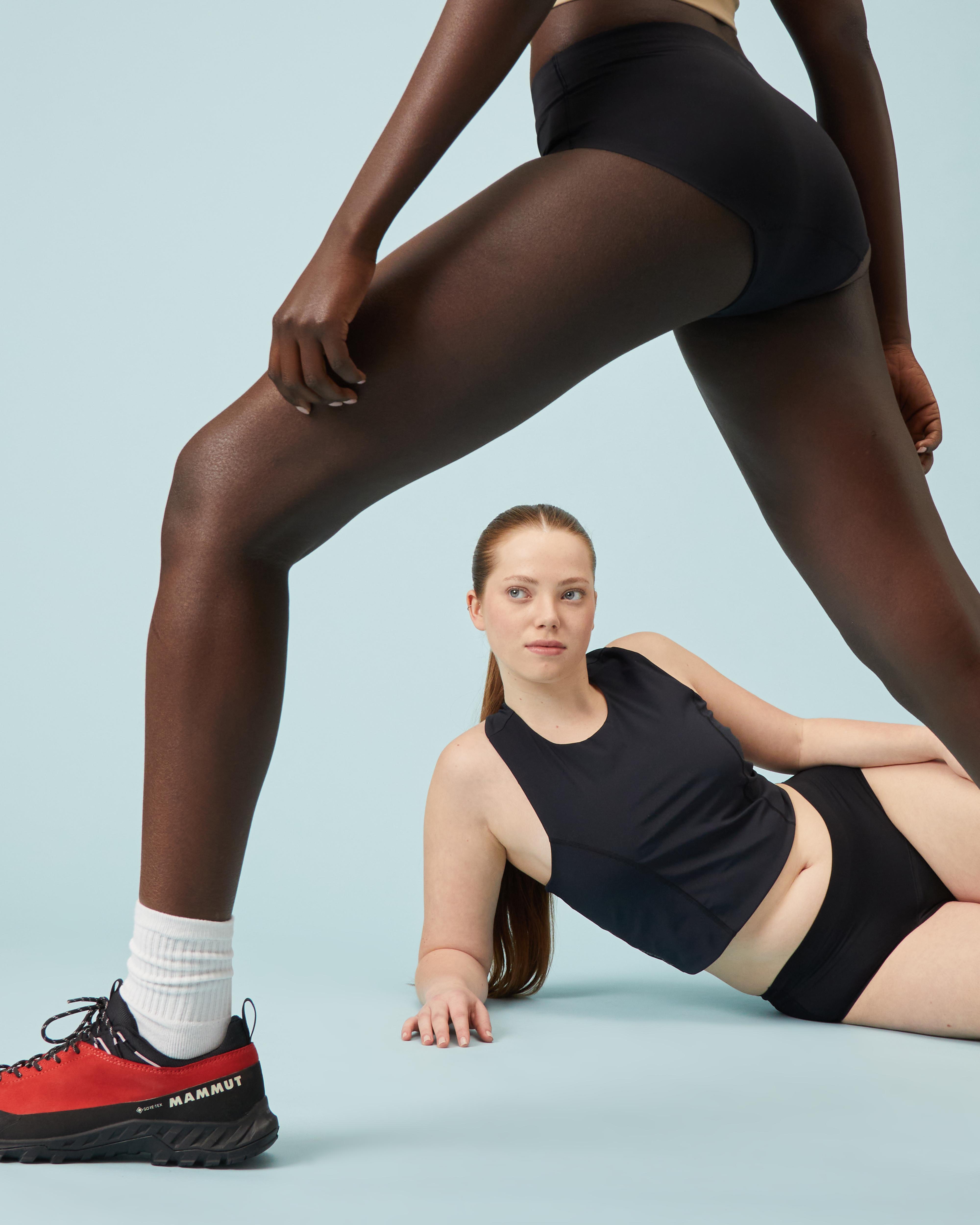 Two women in black Mammut athletic wear pose; one stands with legs apart while the other reclines on the floor between them.