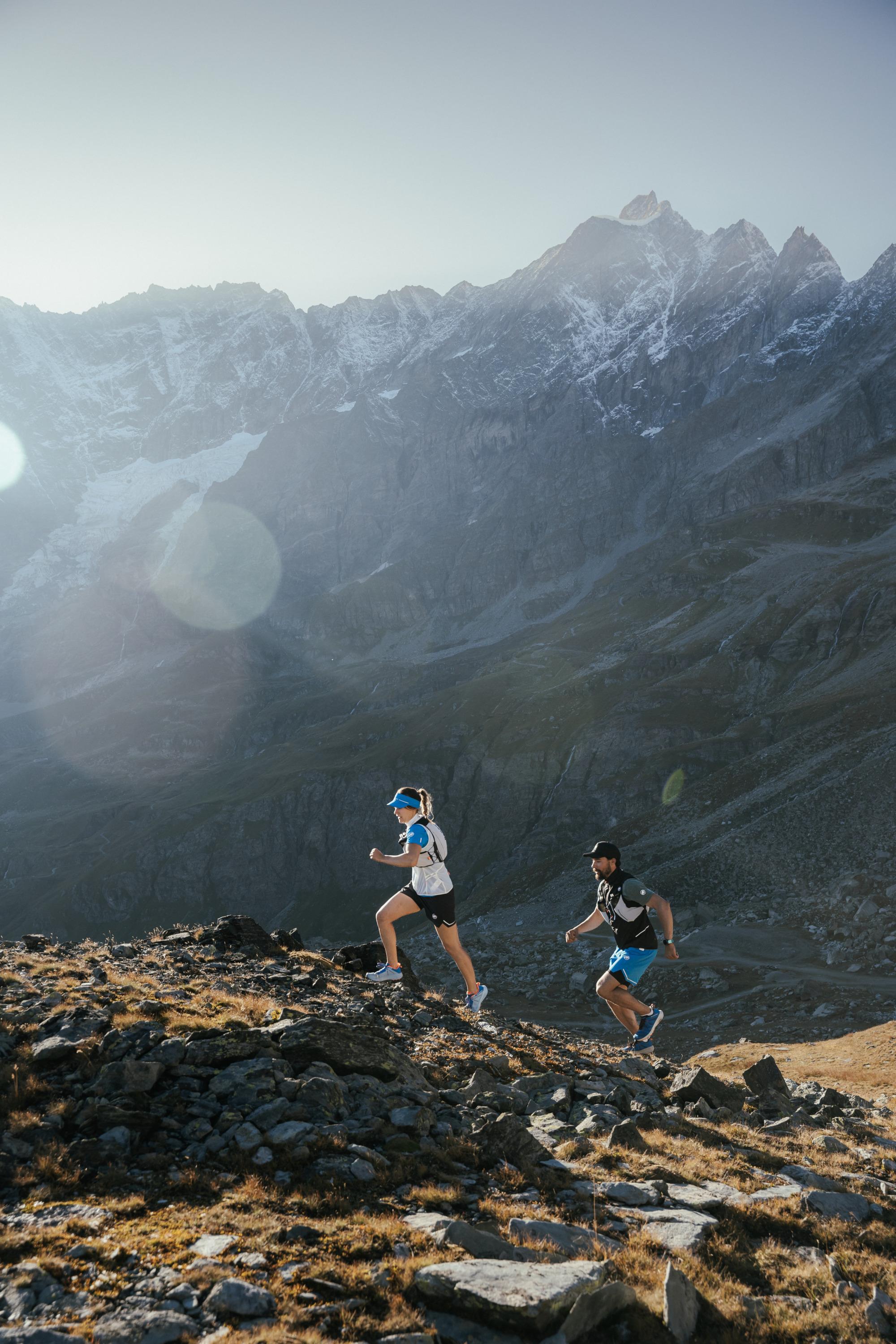 Two trail runners, equipped with Mammut gear, ascend a rugged mountain terrain under a clear sky, with majestic mountains towering in the background.