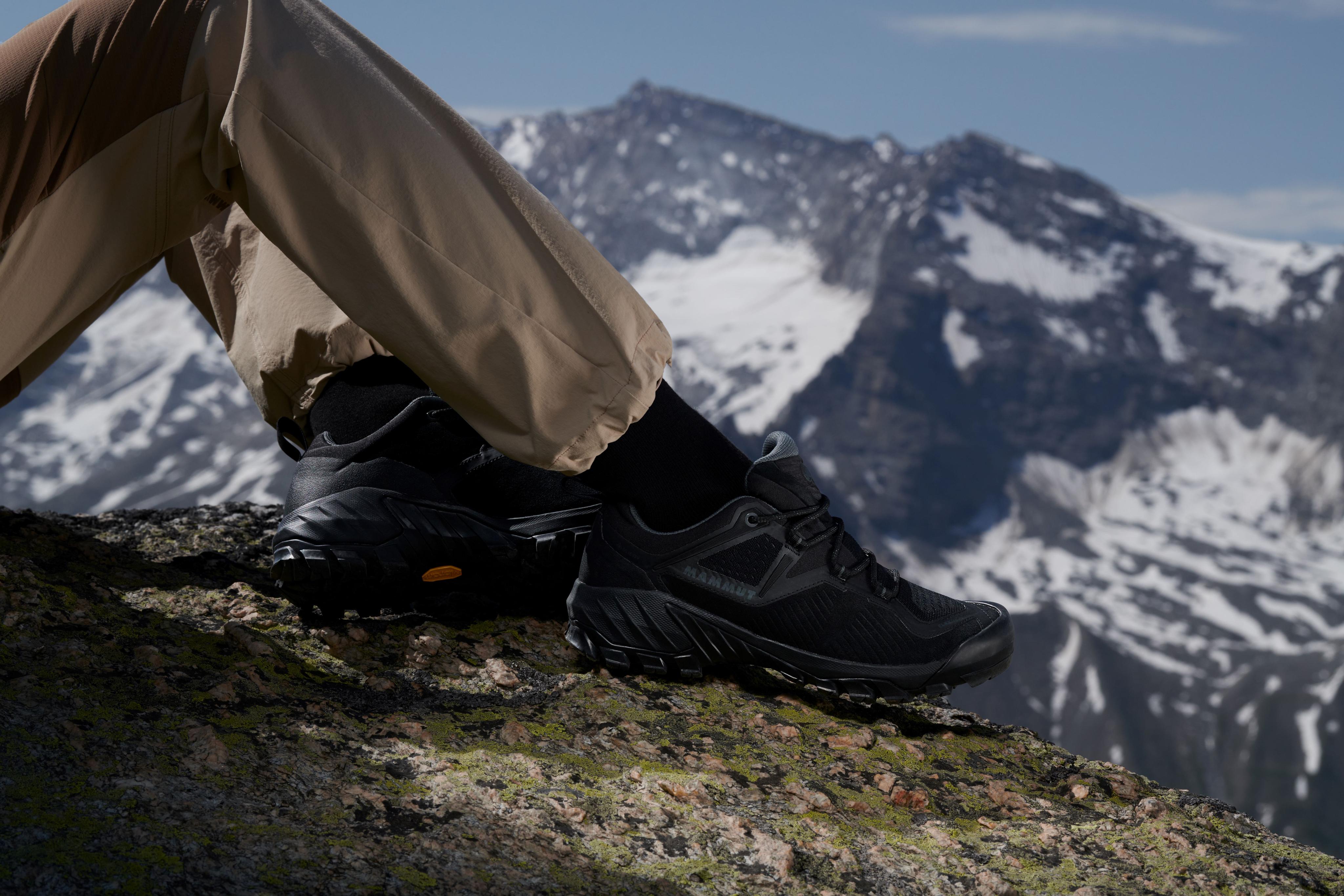Person wearing Mammut black hiking shoes, sitting on a rock with the snowy Alps in the background.