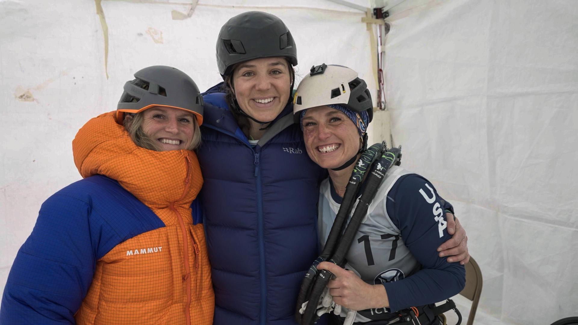 Three smiling women in helmets and Mammut winter gear, posing together inside a white tent during a mountaineering expedition.