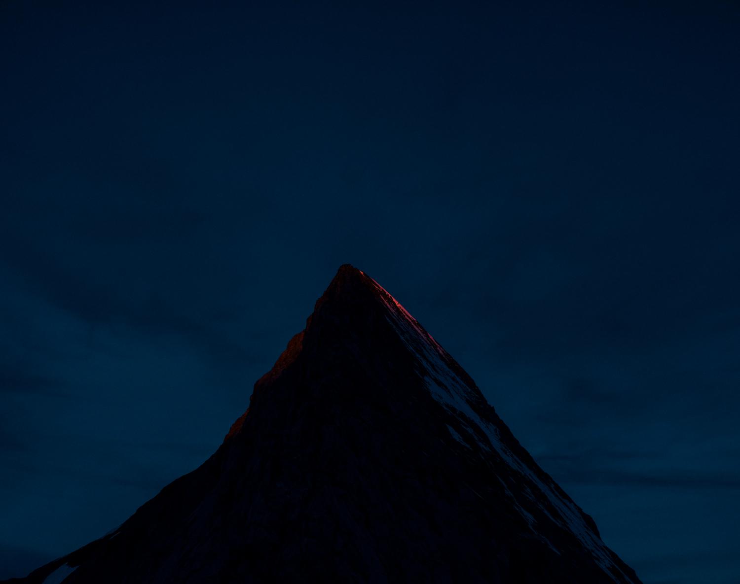 A Mammut-sponsored dark mountain peak is illuminated by a vibrant red glow against a deep blue night sky, highlighting their premium mountaineering gear.