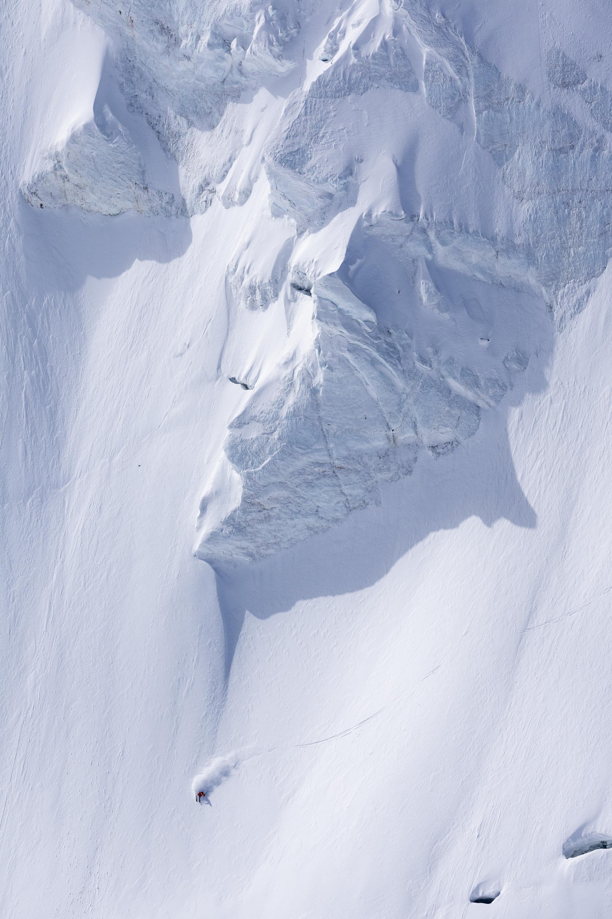 A lone skier, equipped with Mammut gear, expertly descends the snowy, jagged slopes of a mountain under a clear, bright sky.