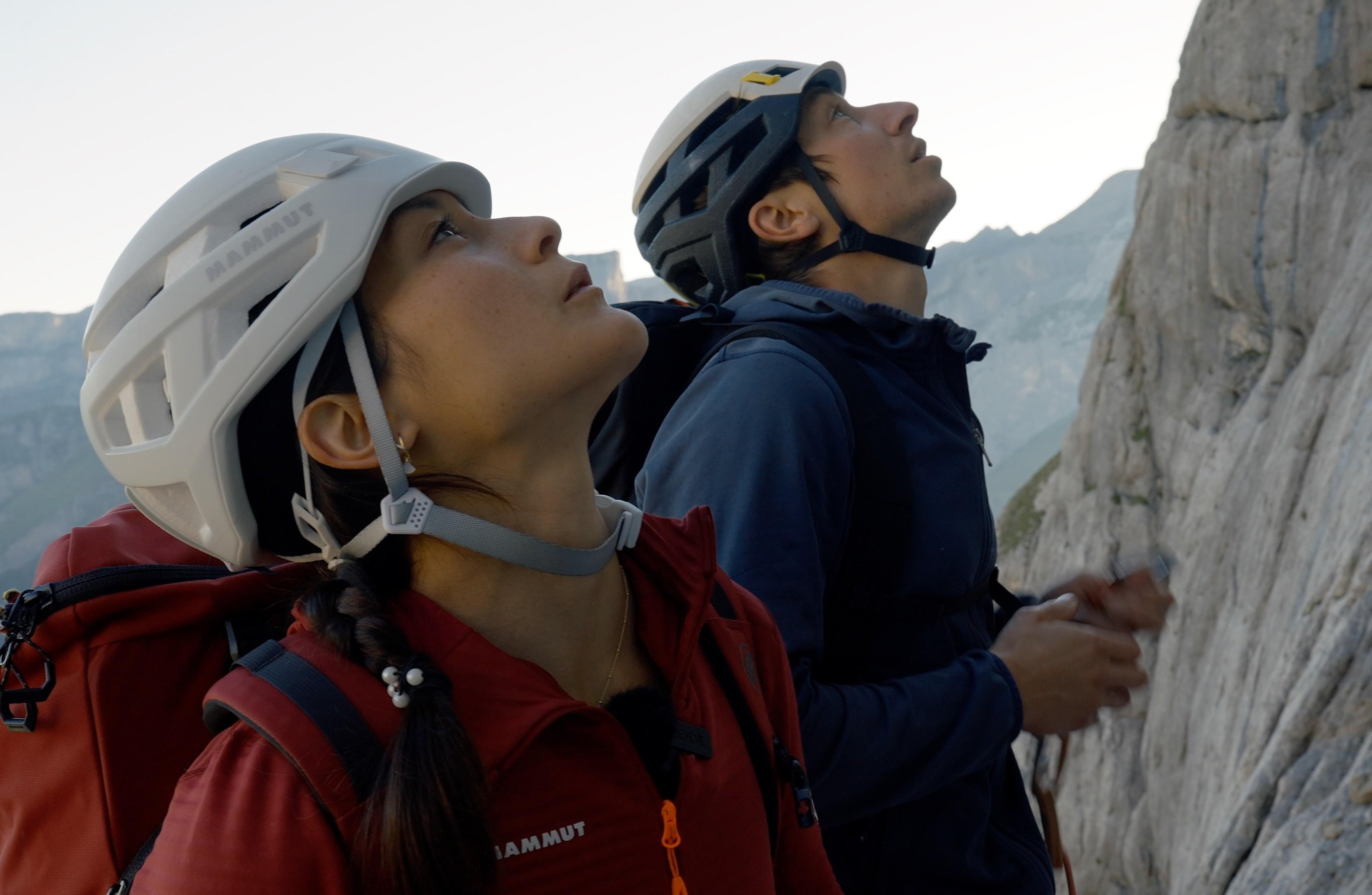 Katharine Choong, a climber wearing Mammut helmets and jackets look up while standing near a rocky cliff face.