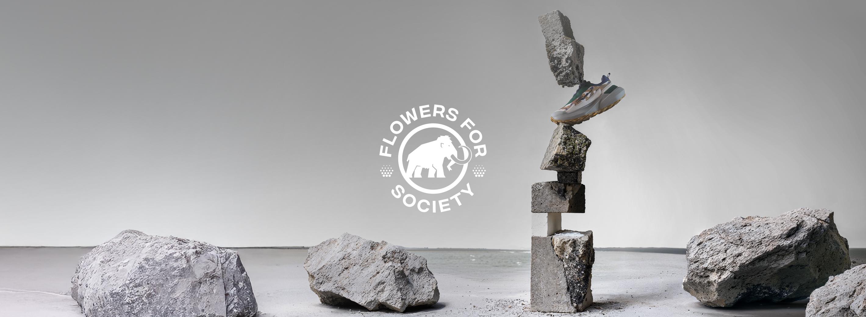 Mammut athletic shoe balanced on stacked concrete blocks, surrounded by large rocks; "Flowers for Society" logo centered, highlighting Mammut's innovative sports footwear design.
