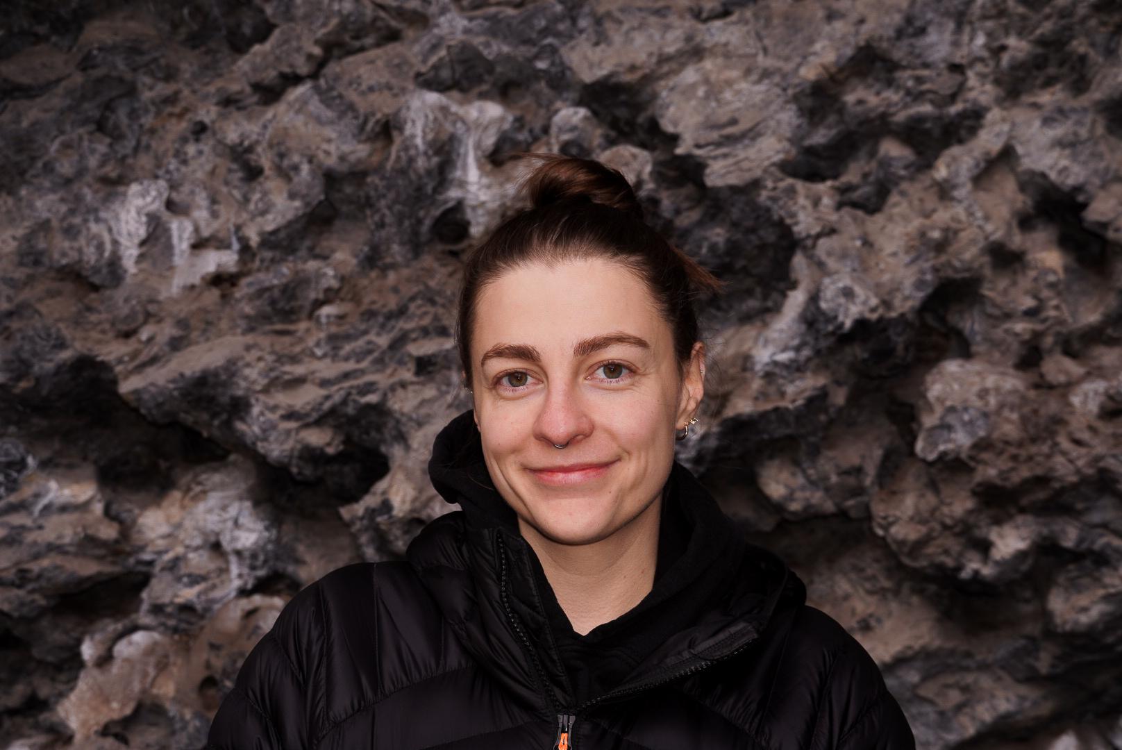A person wearing a Mammut black jacket stands smiling against a rugged rocky background.