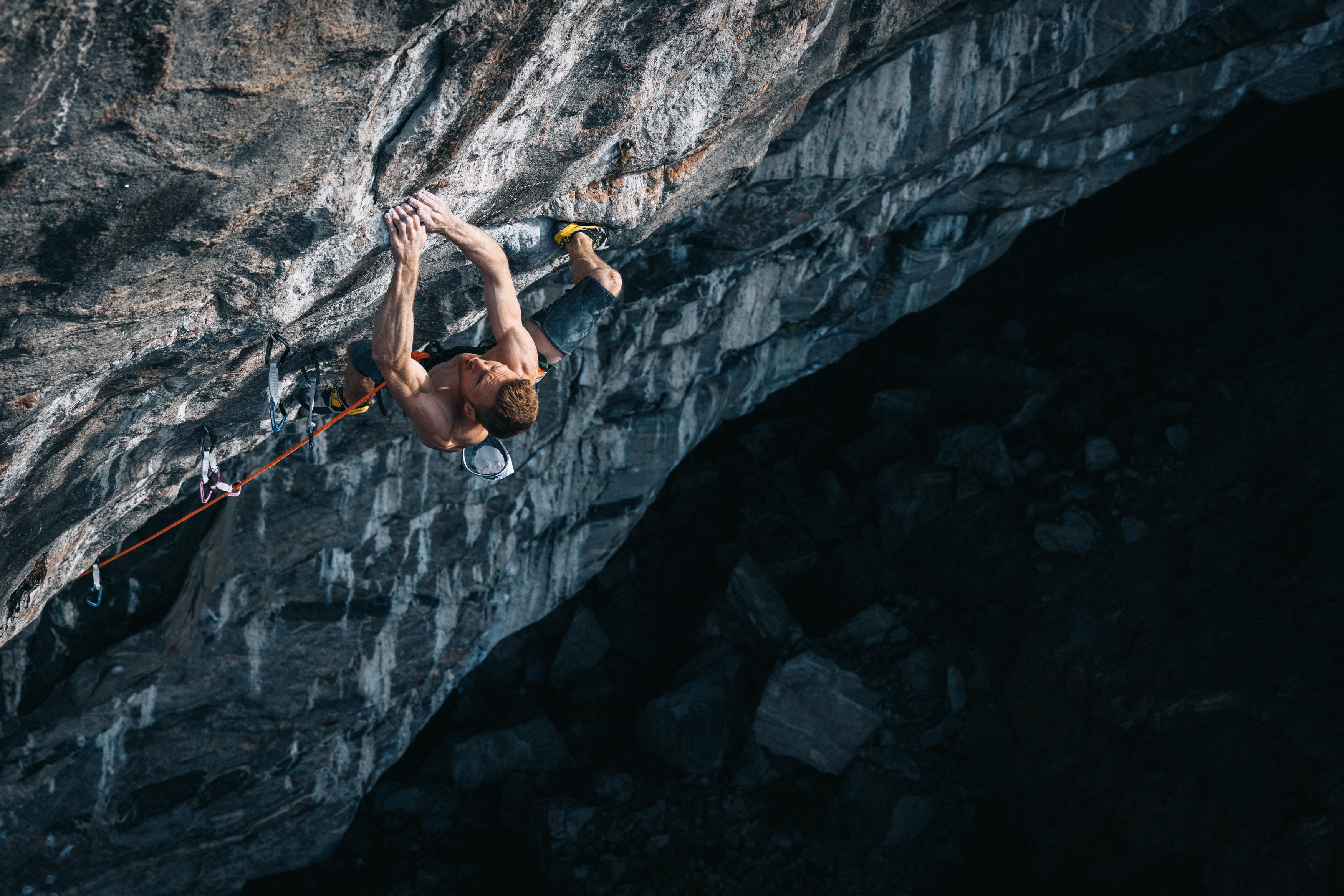 A climber wearing Mammut gear ascends a steep, rocky overhang, gripping the rock face with outstretched arms and legs.