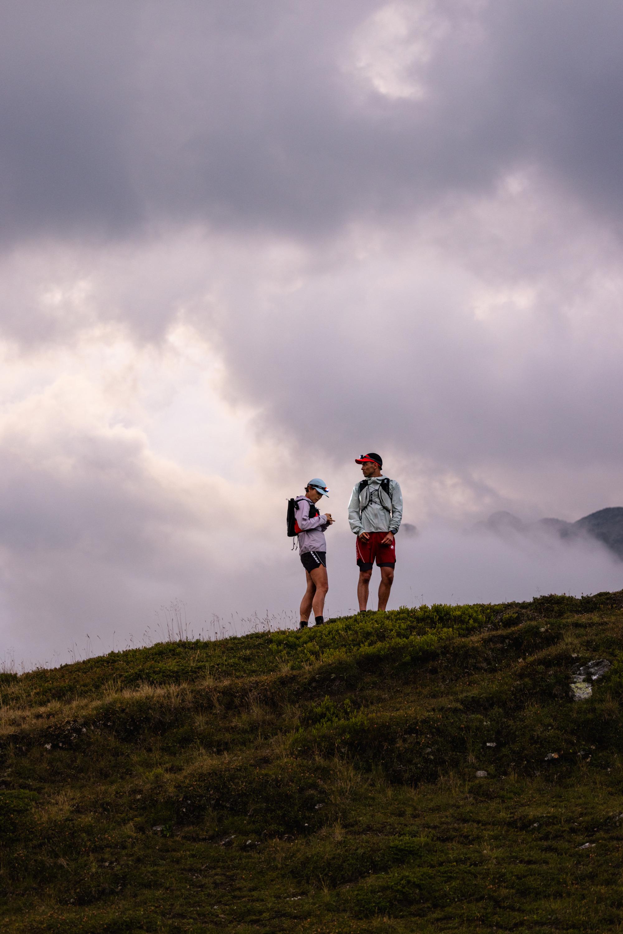 Two hikers in Mammut gear stand atop a rocky peak, overlooking vast mountain ranges at sunset. Ideal moment for adventurers.