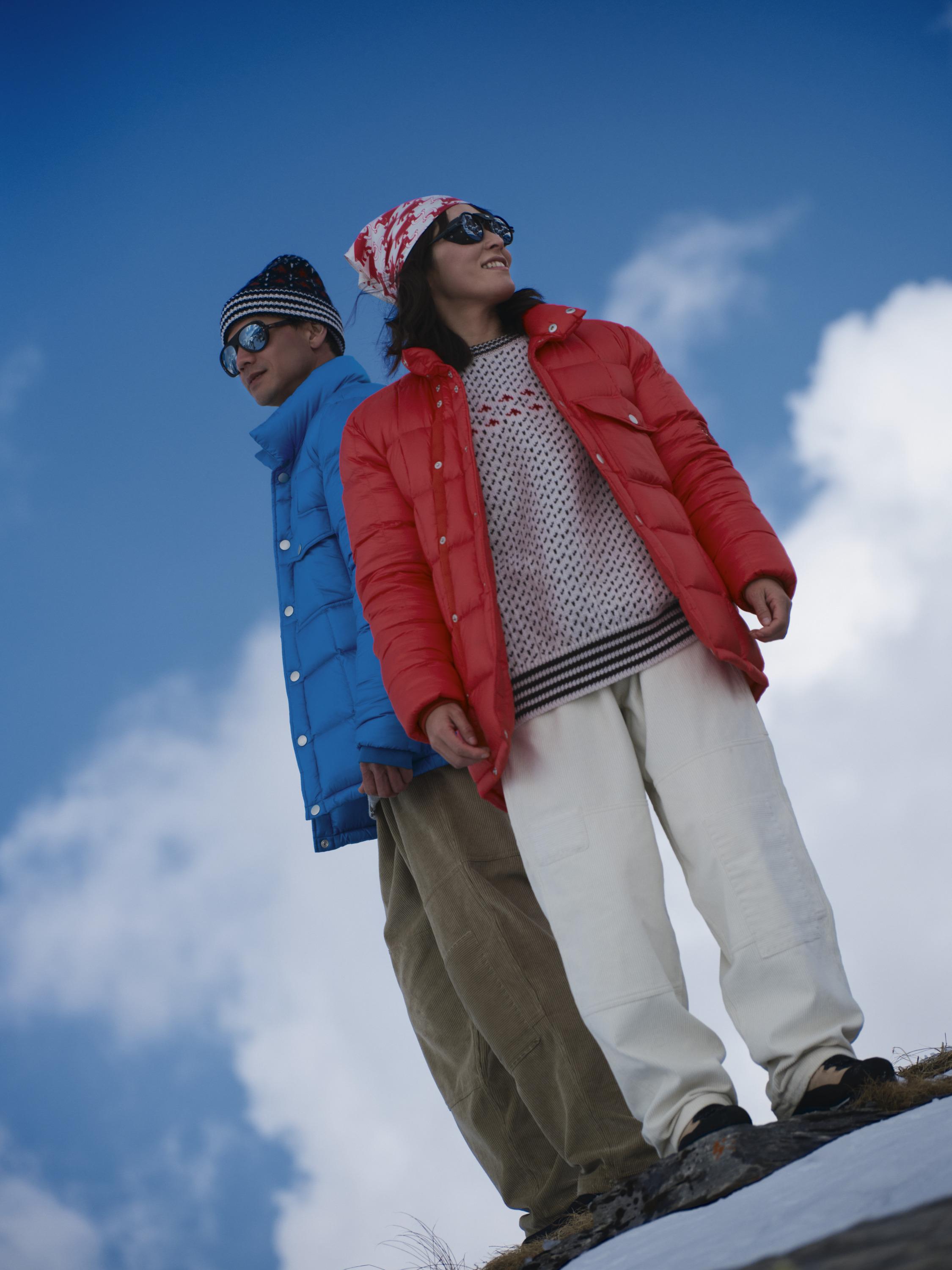 Woman in a purple Mammut jacket stands on a rocky mountain with a lake and hills in the background.