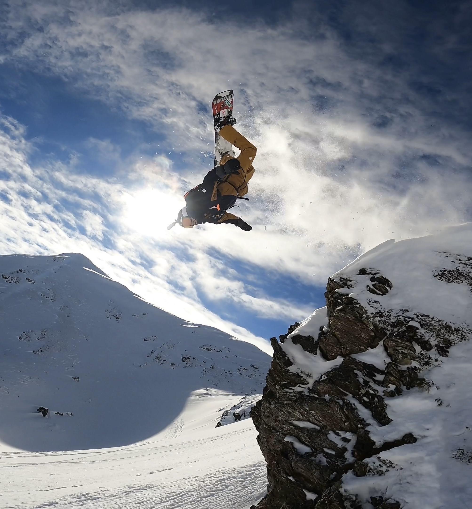 Snowboarder showcasing an aerial trick near a snowy cliff with Mammut gear under a cloudy sky.