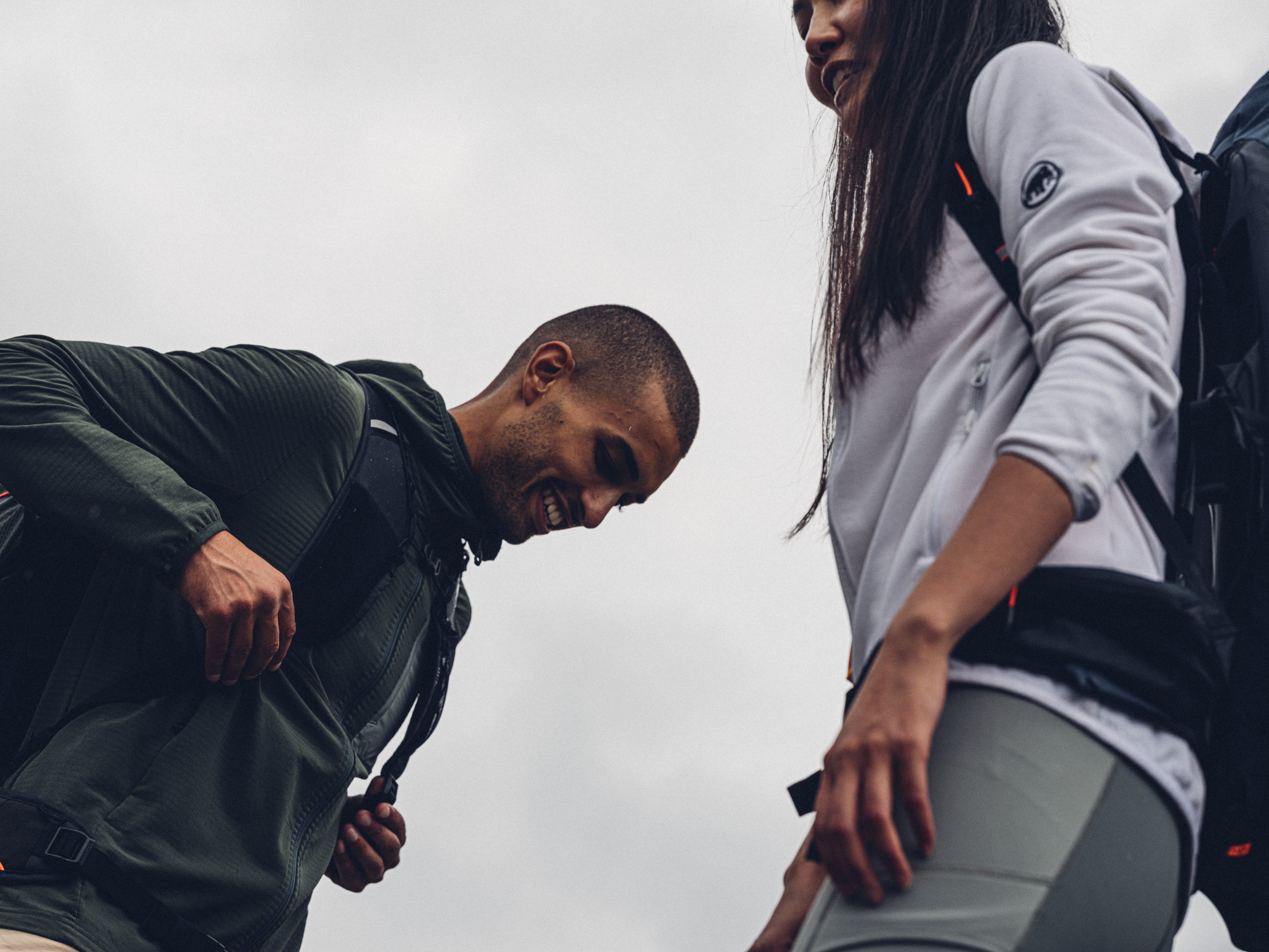 Two people wearing Mammut backpacks smile while hiking outdoors under a cloudy sky, showcasing Mammut outdoor gear.
