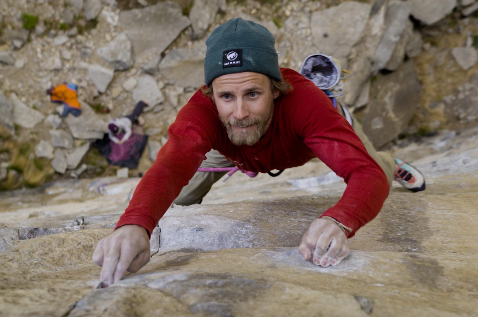 A rock climber in a Mammut red shirt and green beanie ascends a steep cliff, focusing intensely, with chalky hands gripping the rugged rocks.