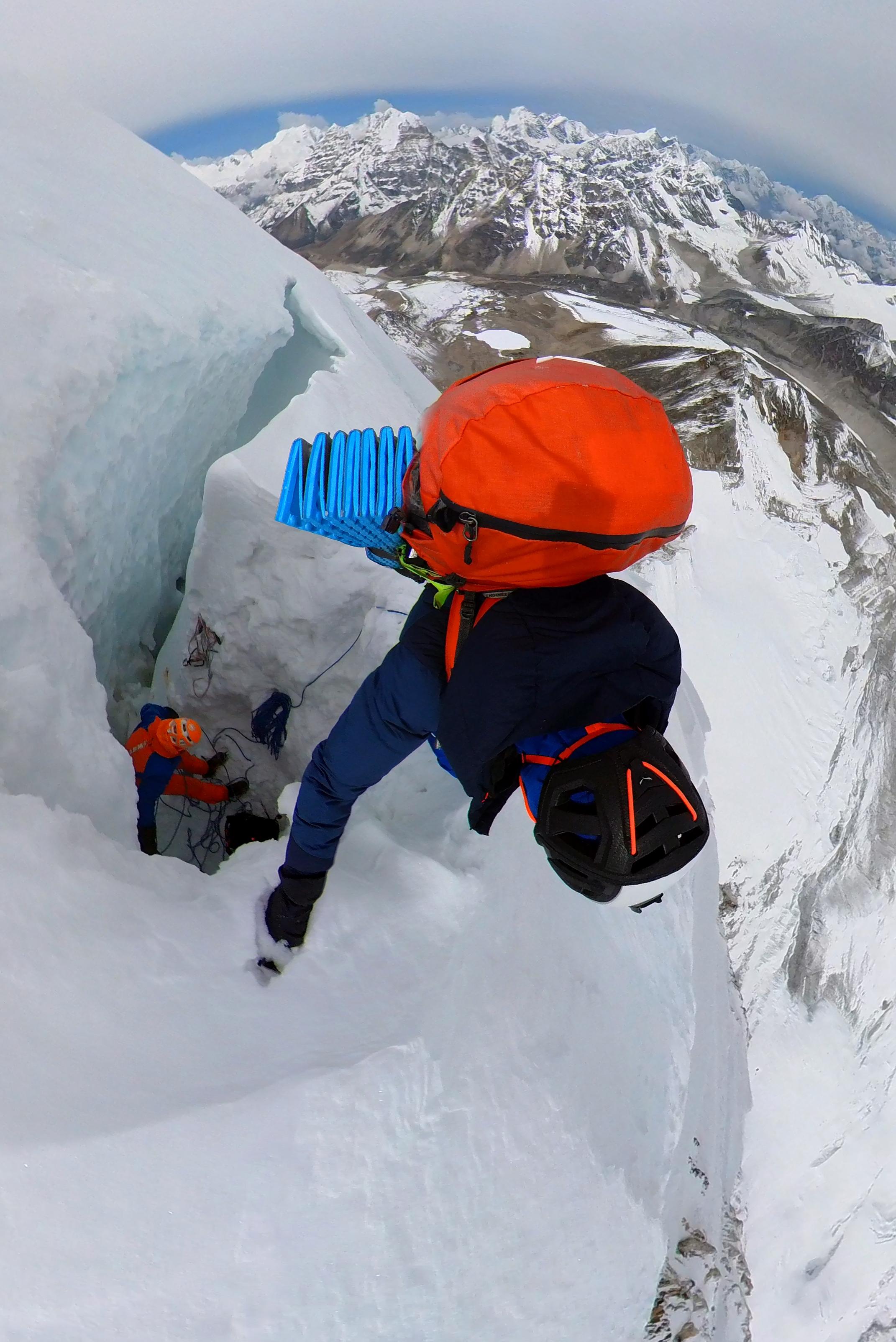 A climber equipped with Mammut gear ascends a snow-covered peak, with another climber following below, set against a stunning backdrop of snow-capped mountains.