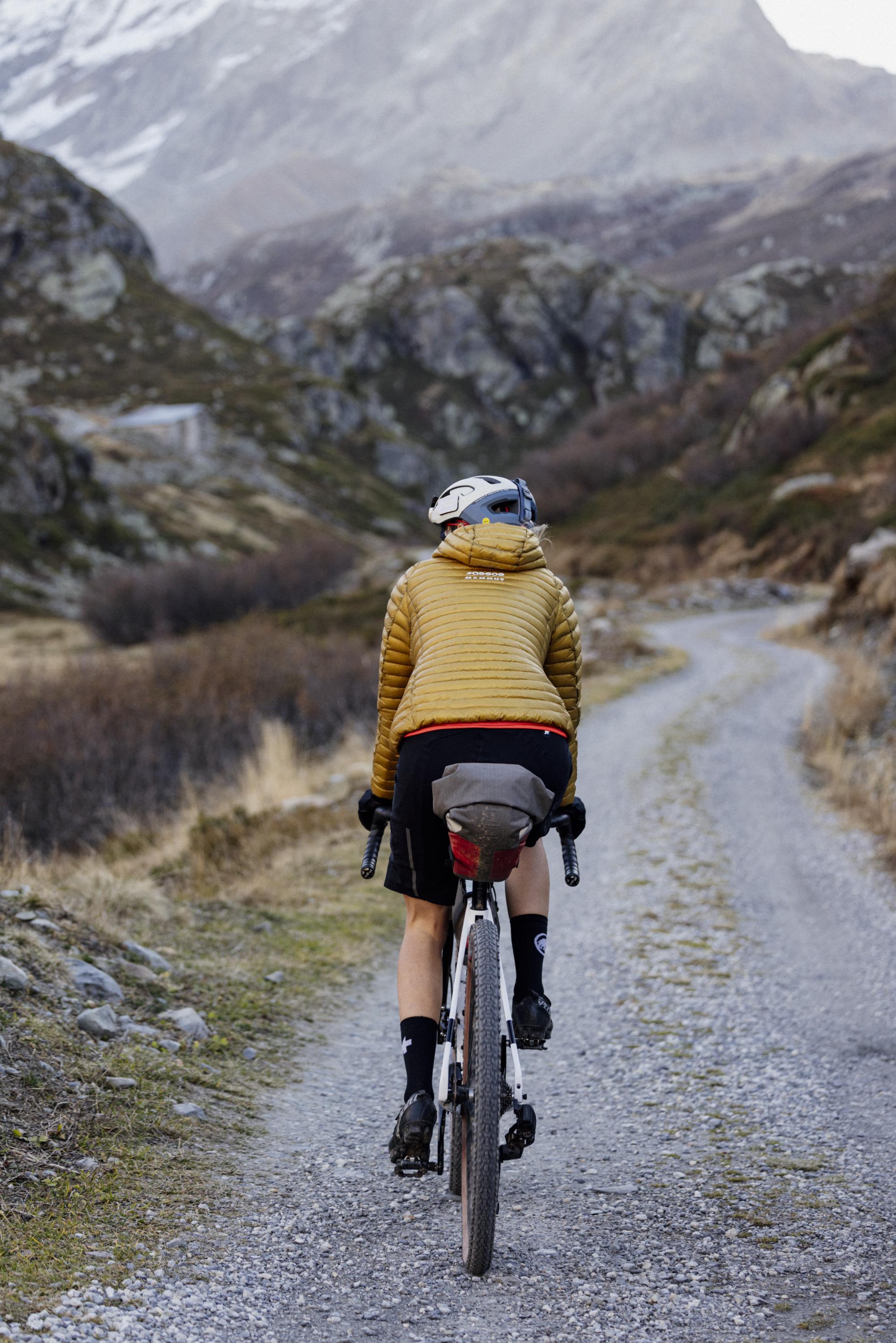 Cyclist on a gravel path through a rugged mountainous landscape, wearing a yellow Mammut jacket and helmet.
