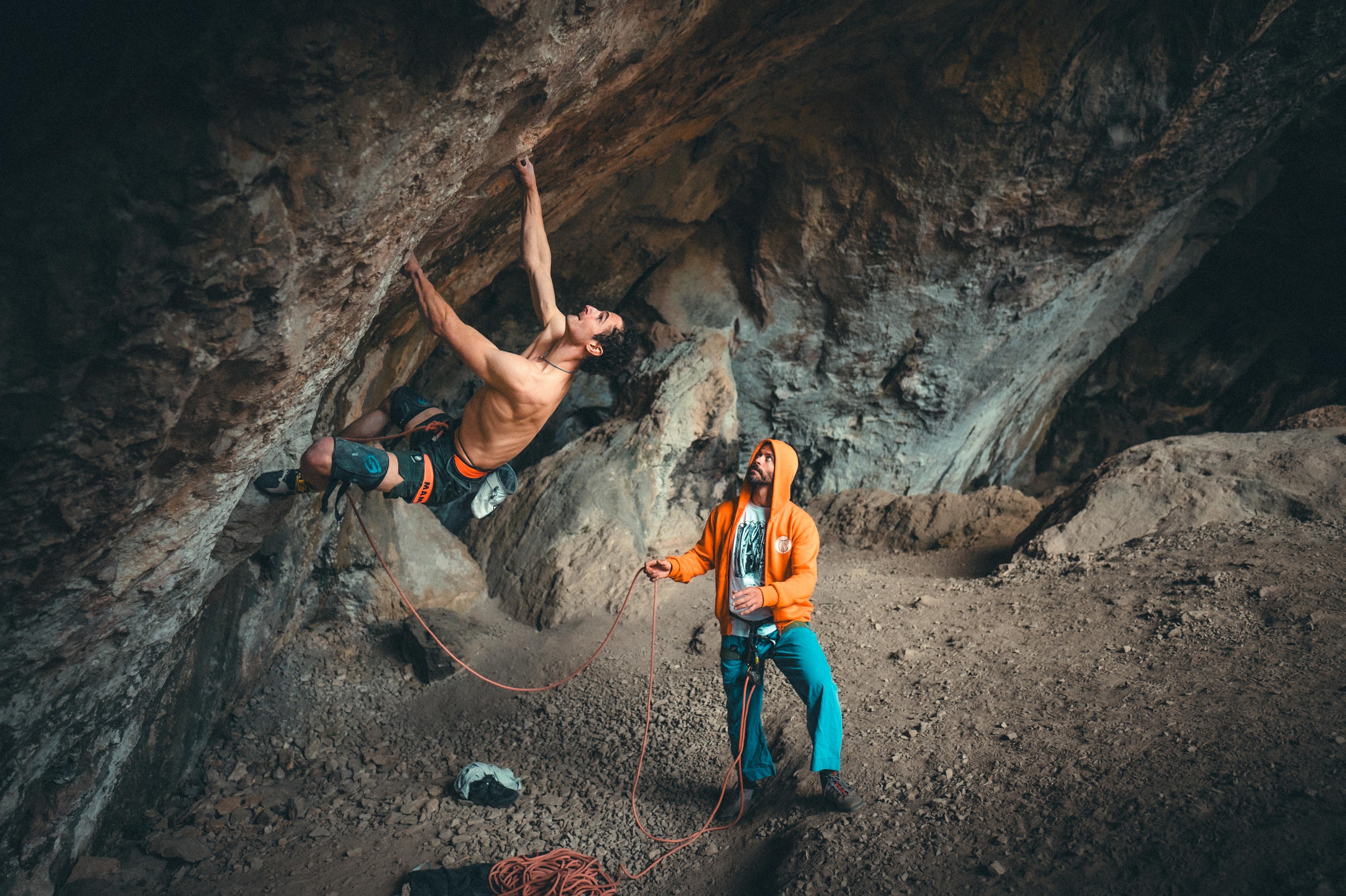 A climber wearing Mammut gear ascends an indoor rock wall while their partner, also outfitted in Mammut climbing equipment, safely belays them from the ground in a cave-like climbing gym.