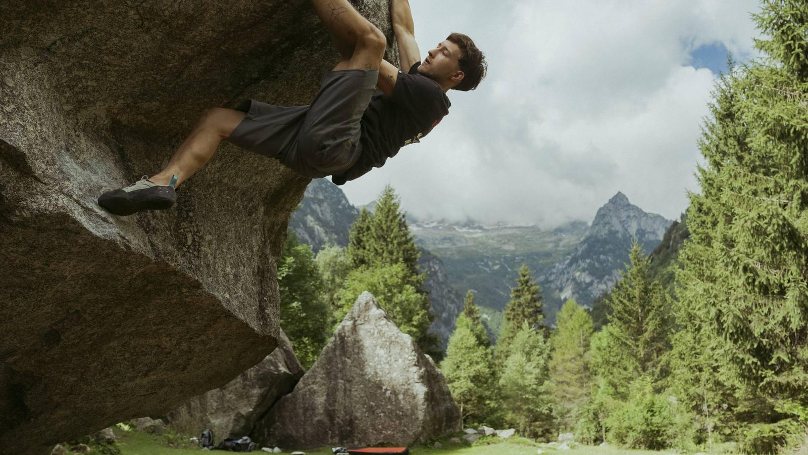 Two Mammut-clad rock climbers, one standing on the other's shoulders, reach for a handhold on a steep rocky cliff face.