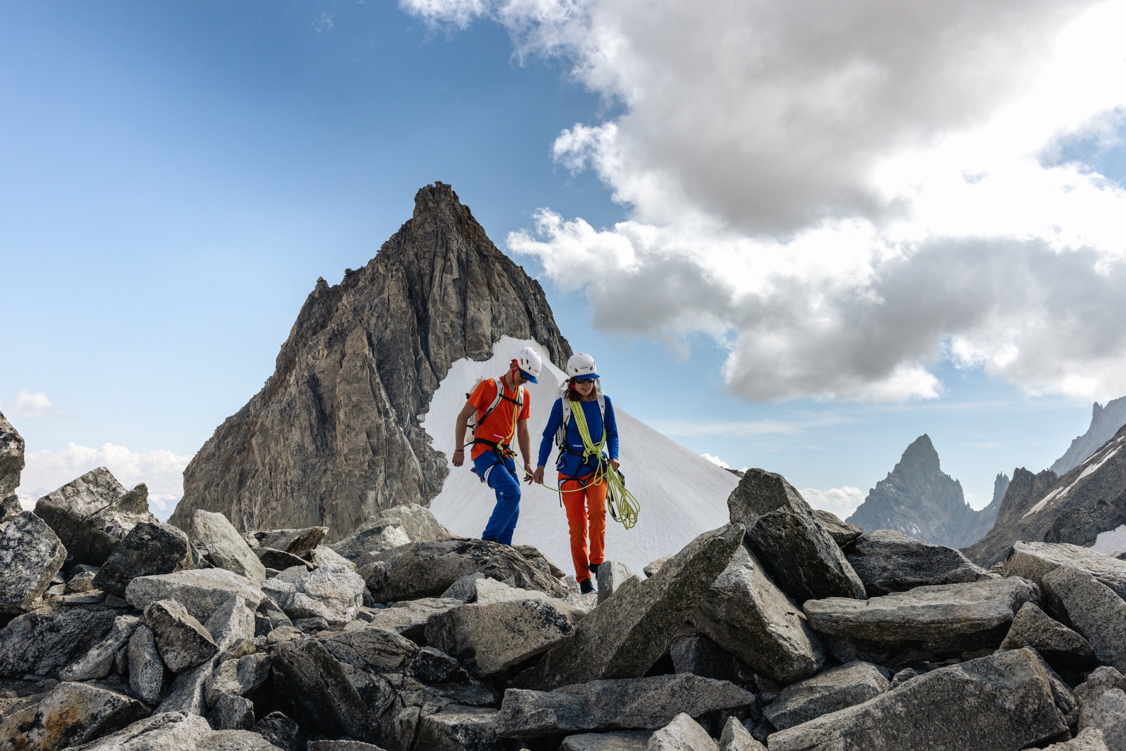 Two climbers wearing helmets and Mammut gear hike over rocky alpine terrain with snowy mountains and clouds in the background.