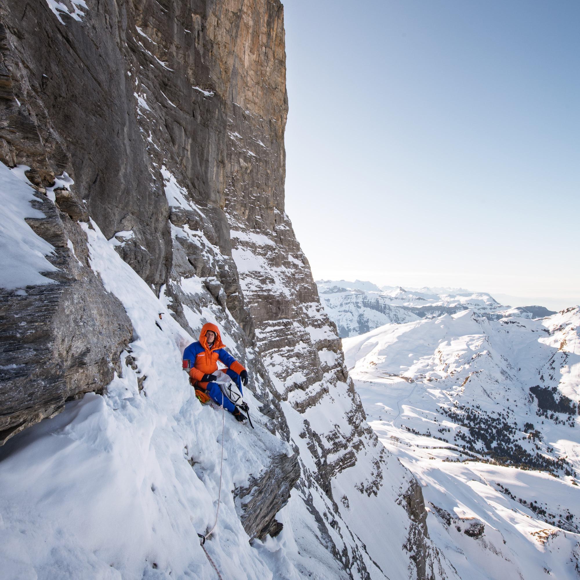 Person in Mammut gear sits on a rocky ledge, gazing over snowy mountain peaks with their legs visible in the foreground.