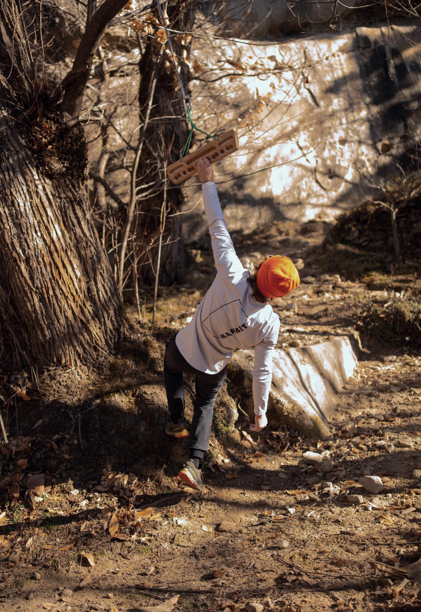 Mammut athlete wearing an orange hat and white Mammut jacket hanging from a rock climbing fingerboard outdoors surrounded by trees.