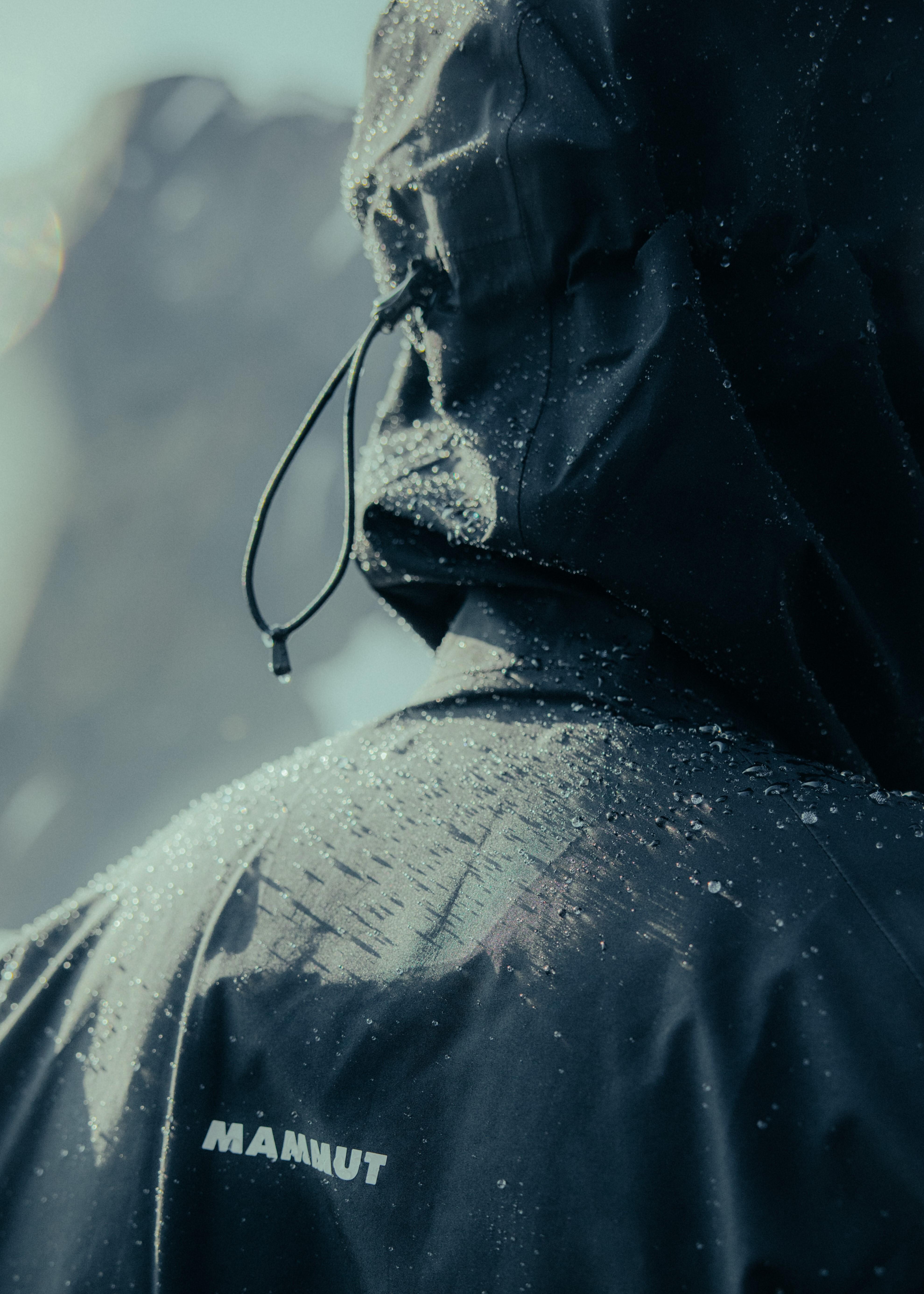 A person in a black Mammut waterproof jacket with a hood, covered in droplets of water, stands facing away from the camera amidst a foggy mountain landscape.