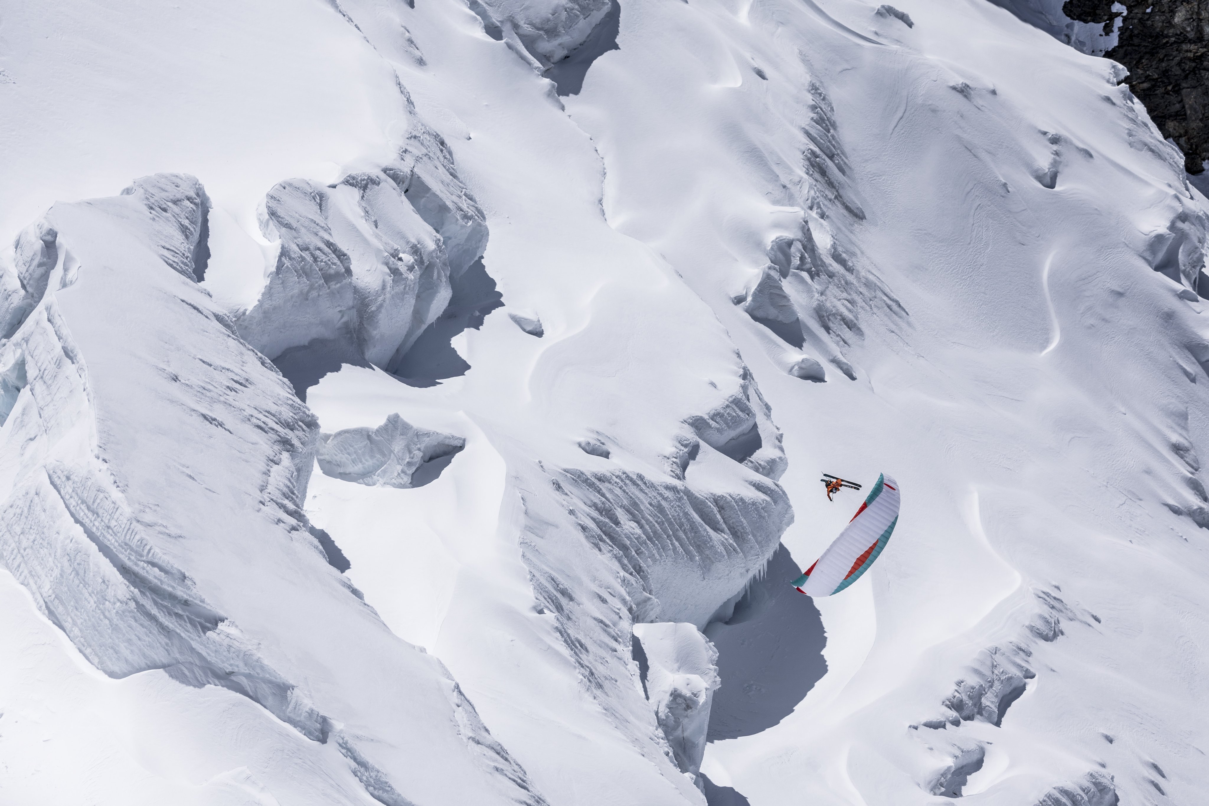 A paraglider wearing Mammut gear navigates snowy, rugged mountainous terrain on a bright day.