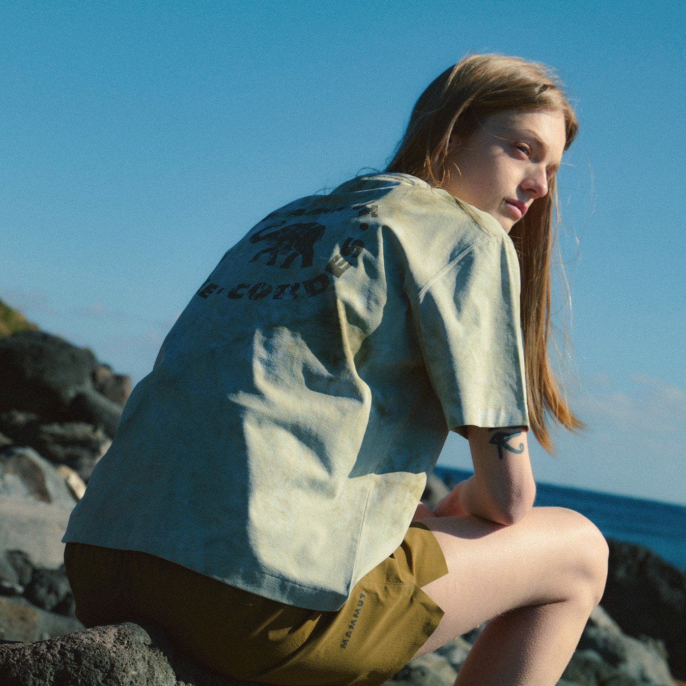 A person with long hair in Mammut activewear sits on seaside rocks, wearing a light t-shirt and brown shorts, looking back.