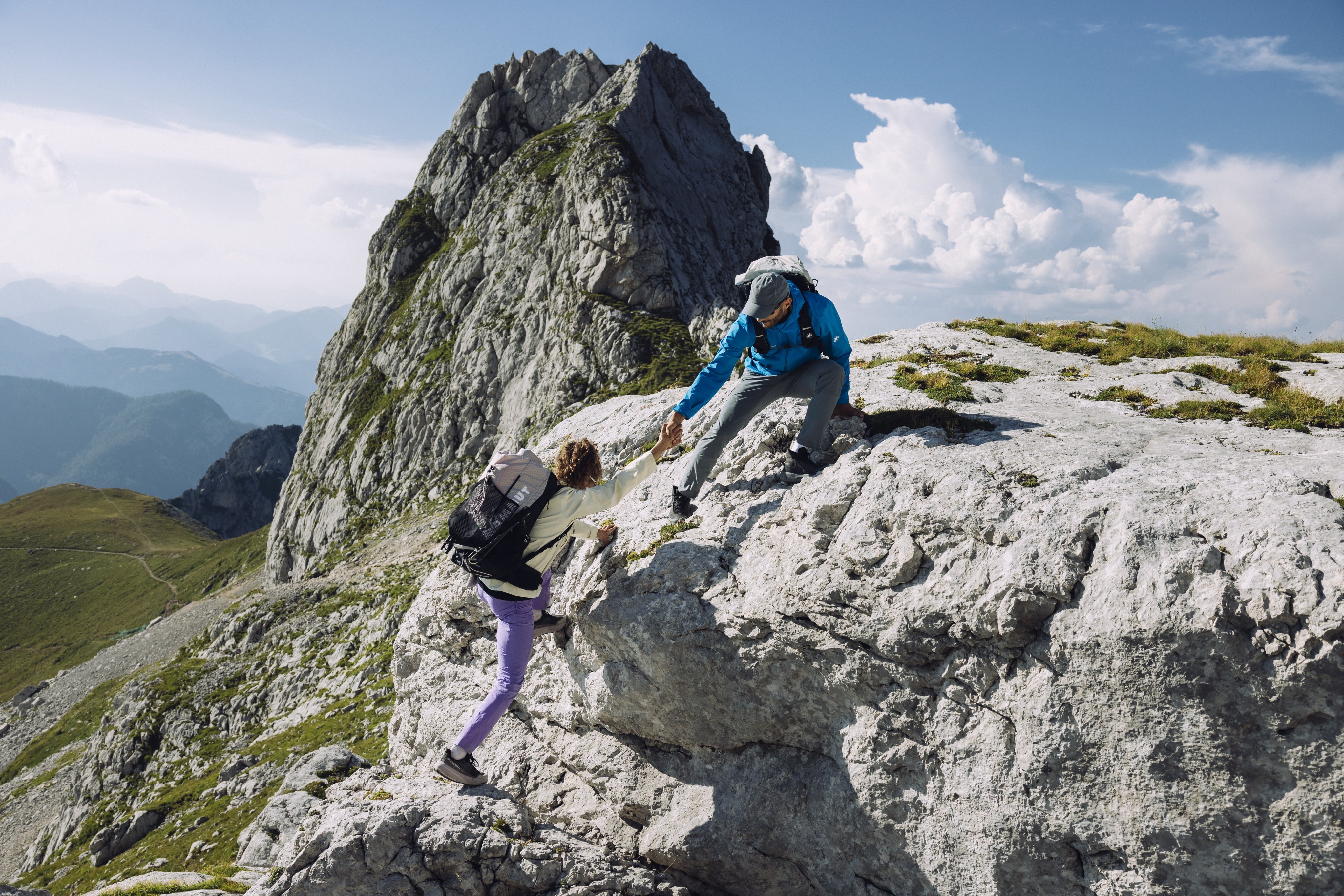 A Mammut-equipped climber assists another in scaling a rocky mountain peak under a clear sky, with distant alpine vistas in the background.