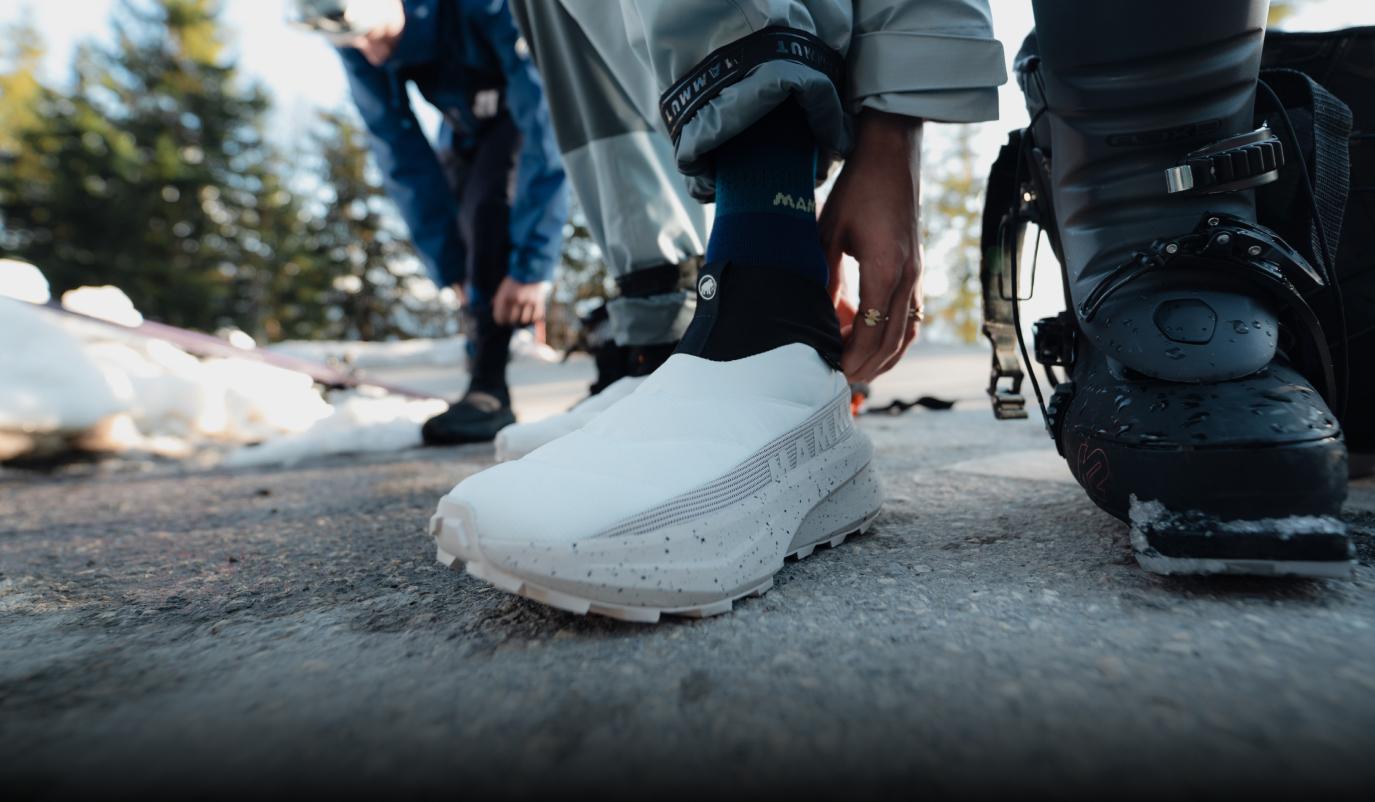 Person adjusts Mammut pant cuffs over white sneakers on snowy ground, wearing dark socks and light-colored pants.