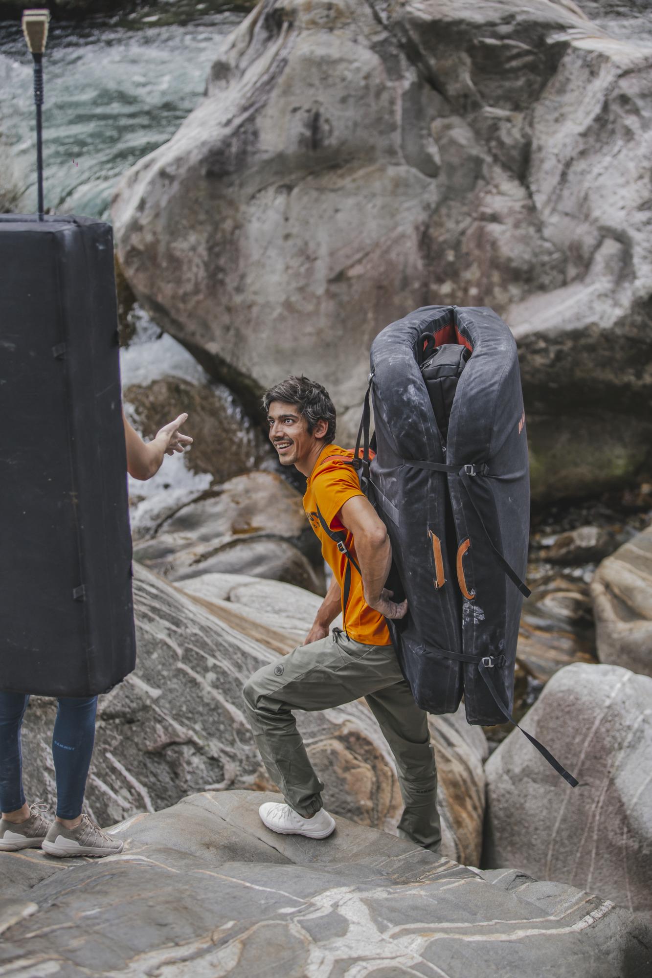 Man in Mammut orange shirt carrying a large black climbing pad, navigating rocky terrain near a river. Another person points ahead, guiding the climb.