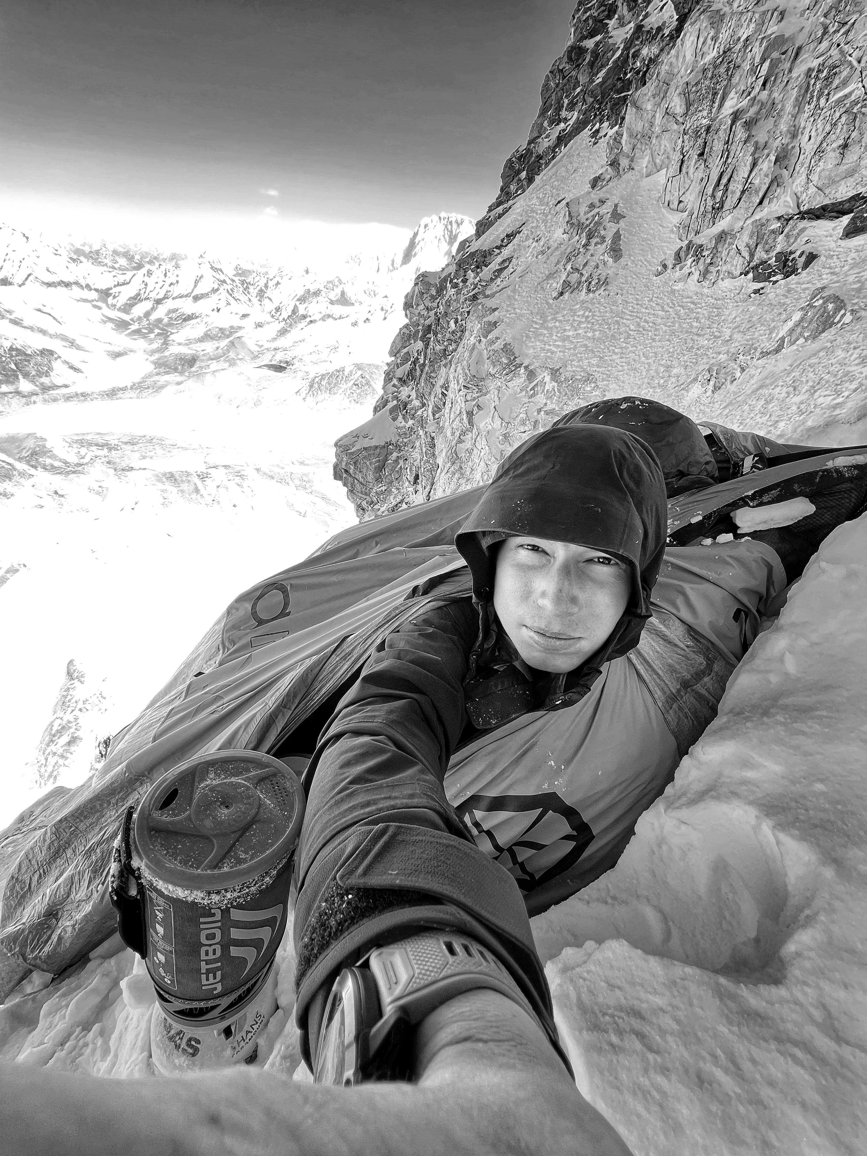 A climber in a Mammut hooded jacket takes a selfie on a snowy mountain ledge next to a steep rock face.