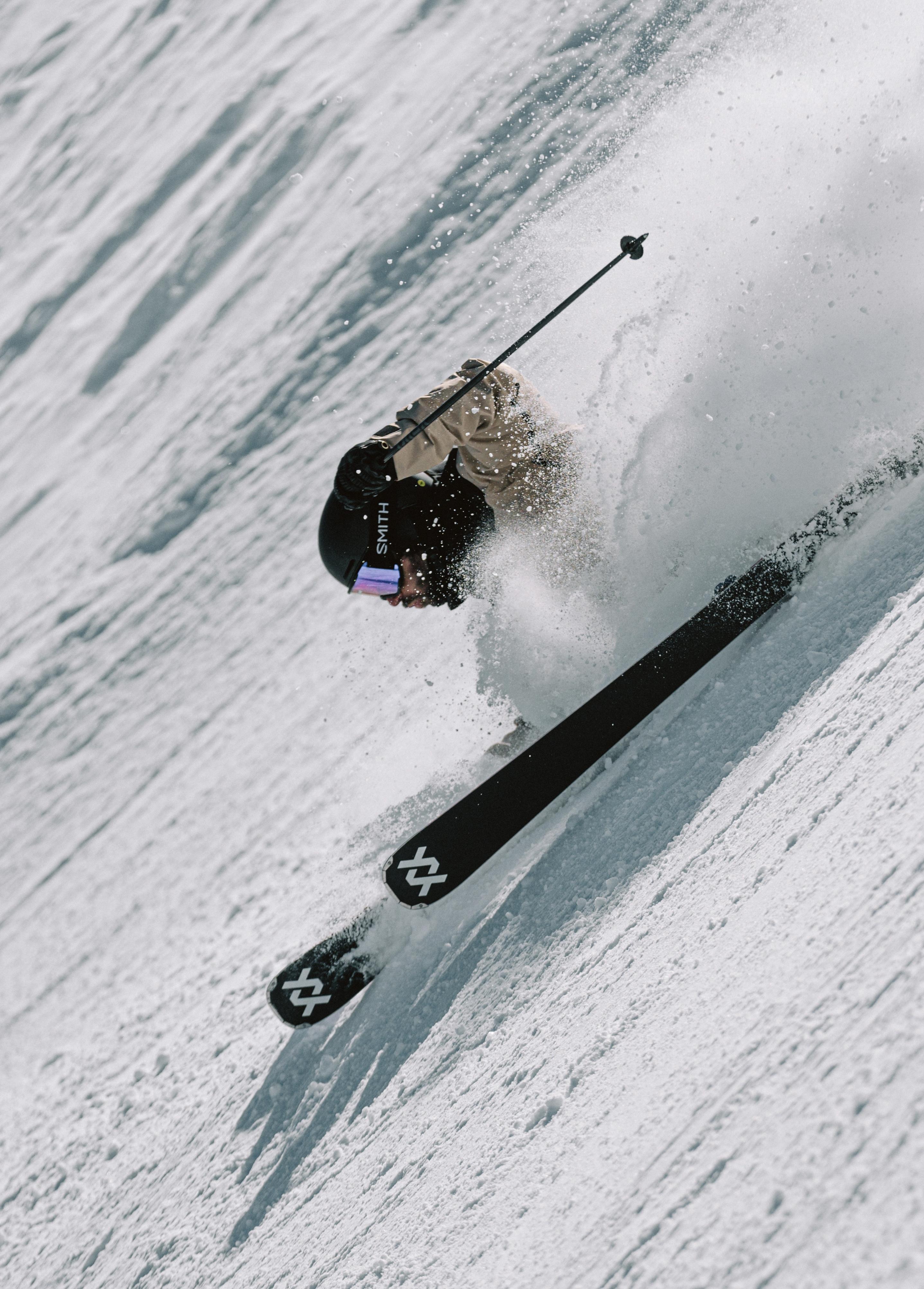 Mammut skier in beige jacket and black helmet speeding down a snowy slope, kicking up powder while showcasing Mammut winter sports gear.