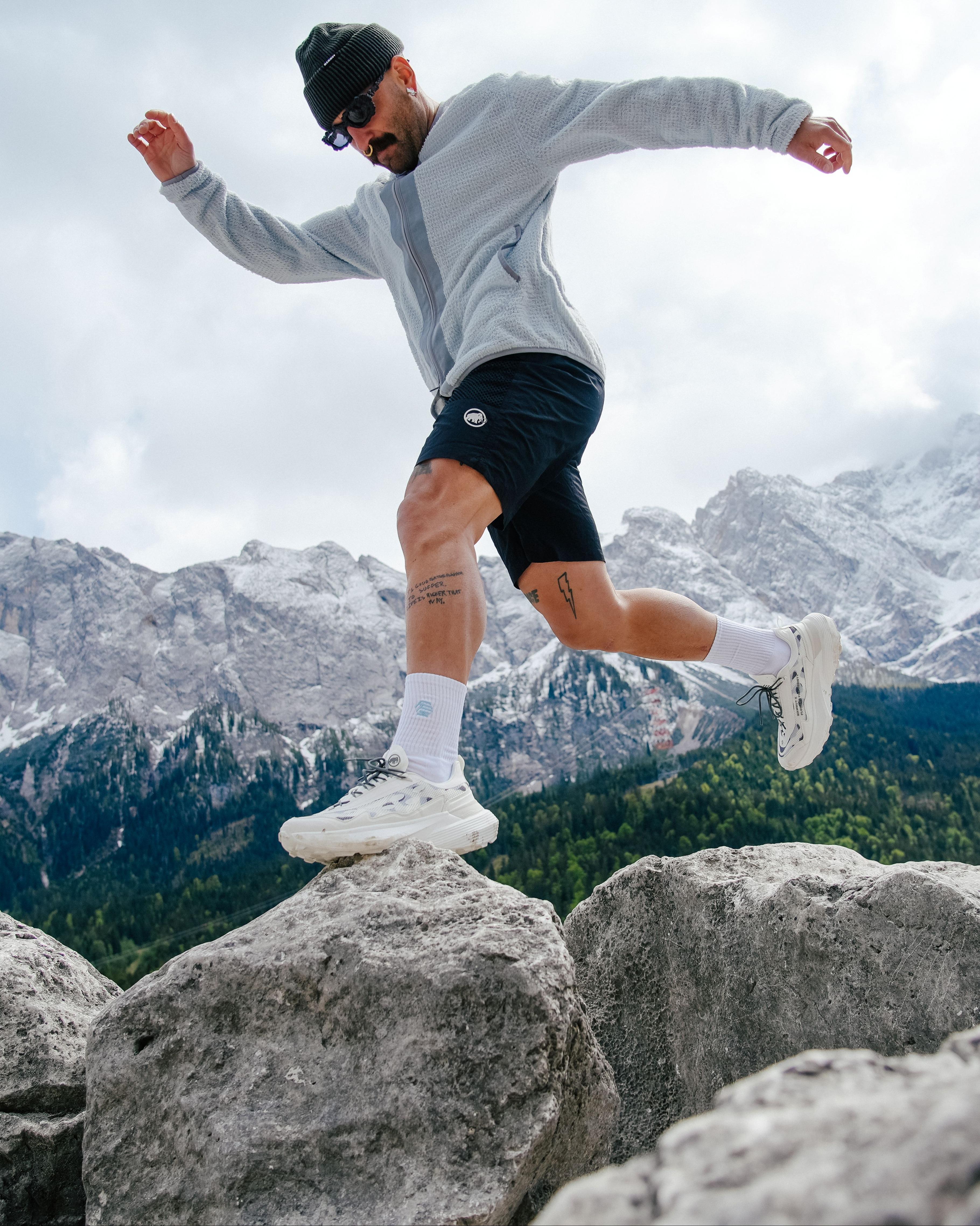 Man in Mammut athletic wear leaps between rocky boulders with snowy mountain peaks and dense forest visible in the background.
