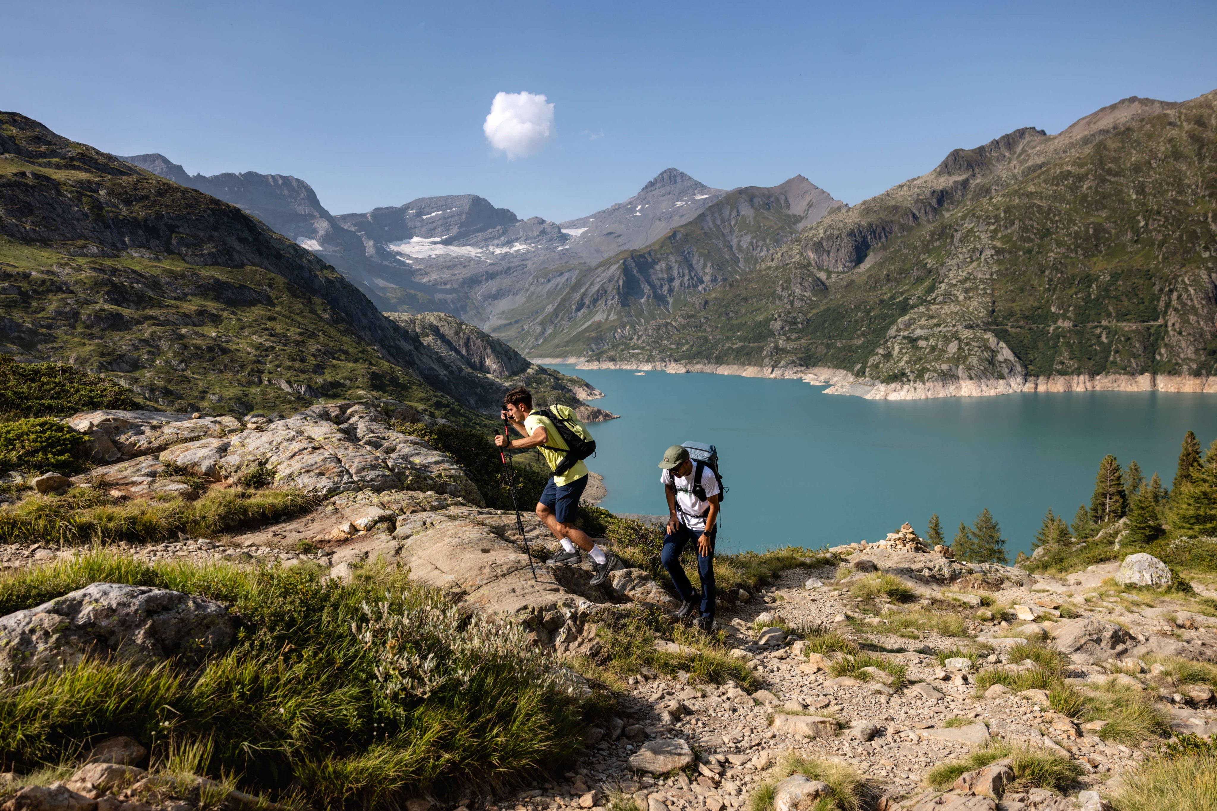 Two hikers in Mammut backpacks trek uphill on a rocky mountain trail above a blue lake and peaks under a clear sky.