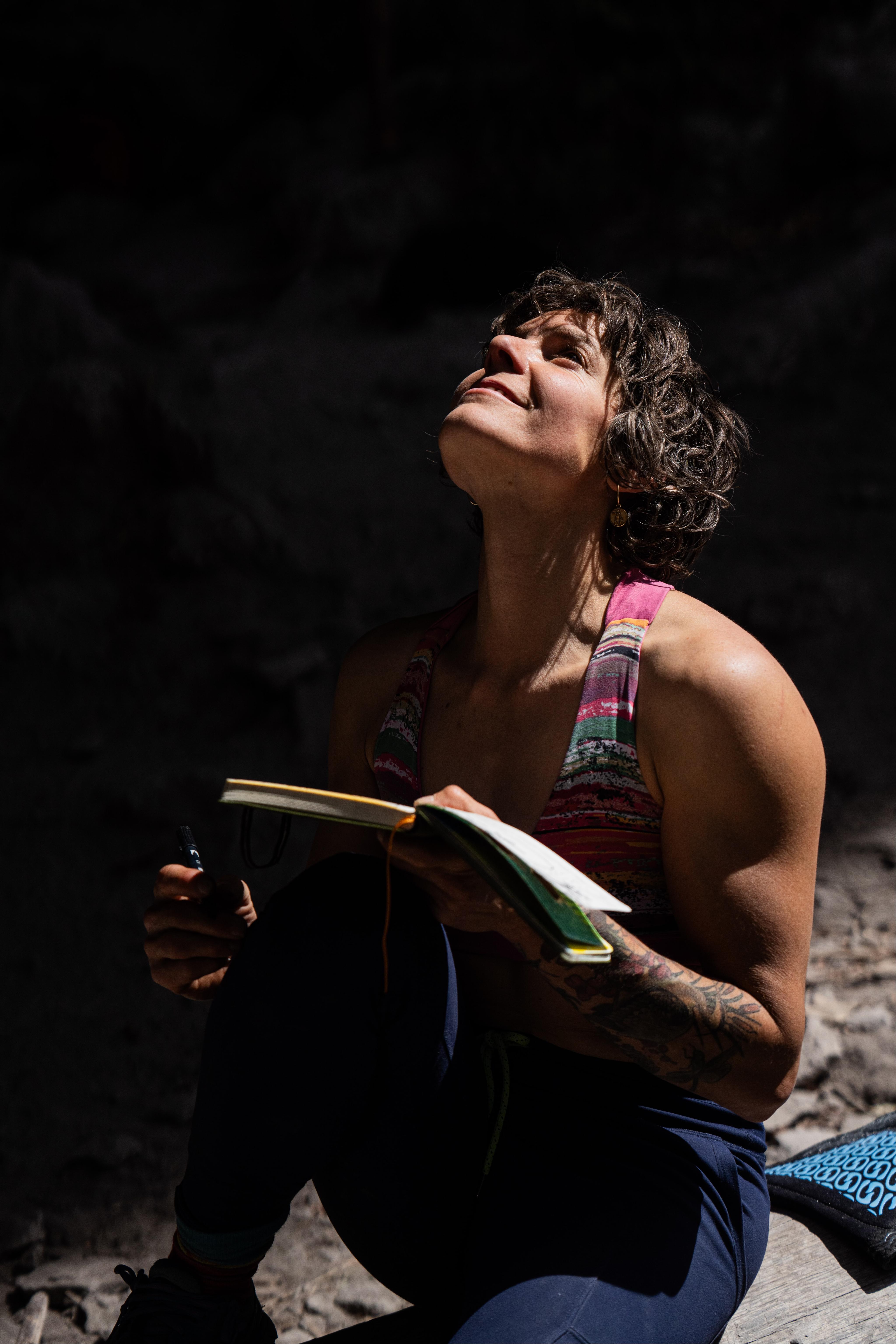 Katie Lambert in a Mammut sleeveless top sits outdoors, thoughtfully gazing upwards while holding a journal and pen.