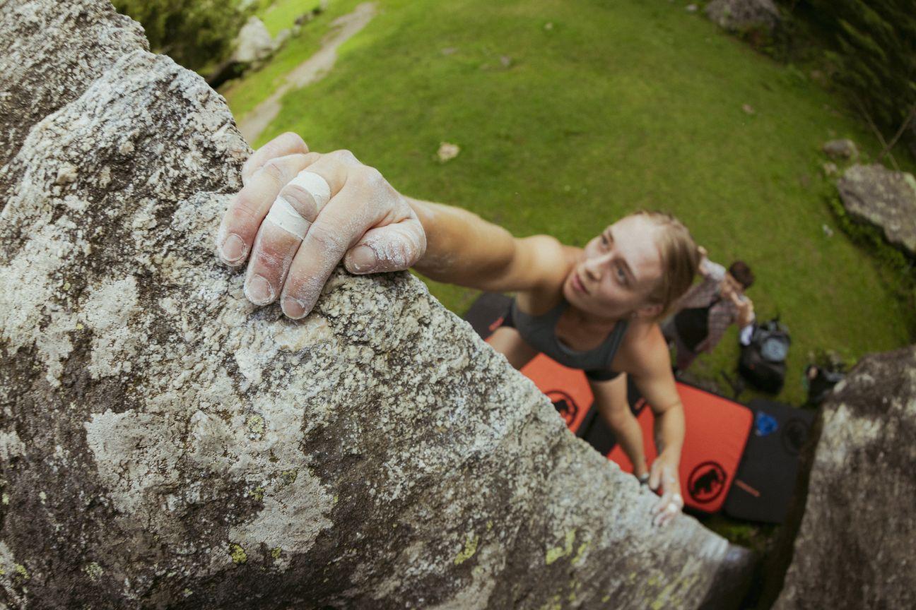 A climber in a helmet ties his Mammut climbing shoes, with Mammut rope and gear nearby, beside a rugged outdoor rock wall.