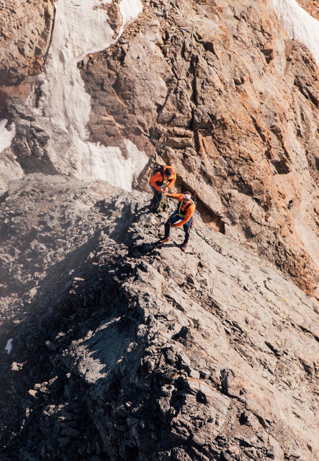 Two climbers wearing orange Mammut jackets on a rugged, rocky mountain ridge, surrounded by patches of snow.