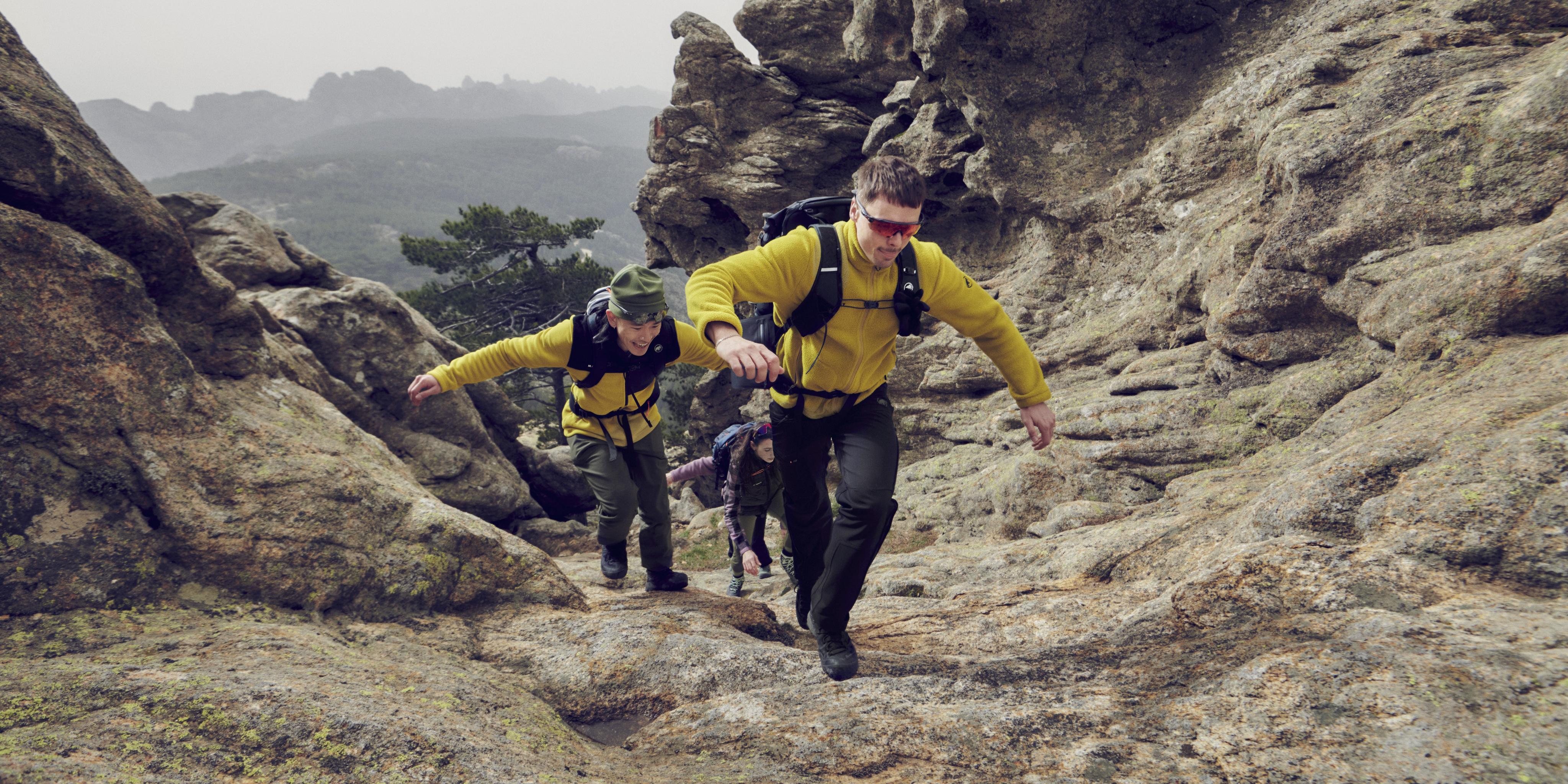 Two hikers in yellow Mammut jackets ascend a rocky mountain trail, showcasing a breathtaking view of distant peaks in the background.