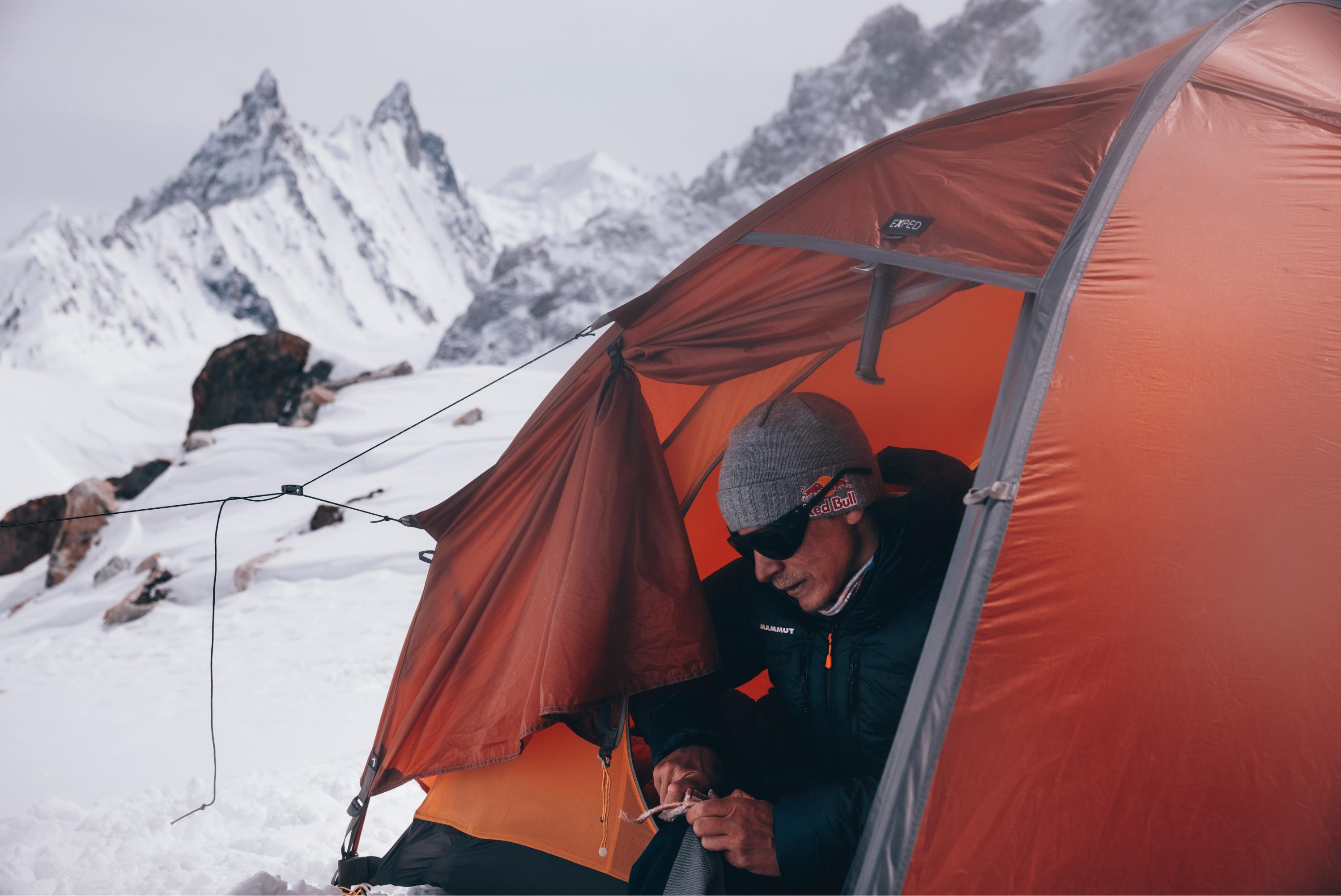 Person in a beanie and sunglasses inside a Mammut orange tent, set up in a snowy mountain landscape with majestic peaks in the background.